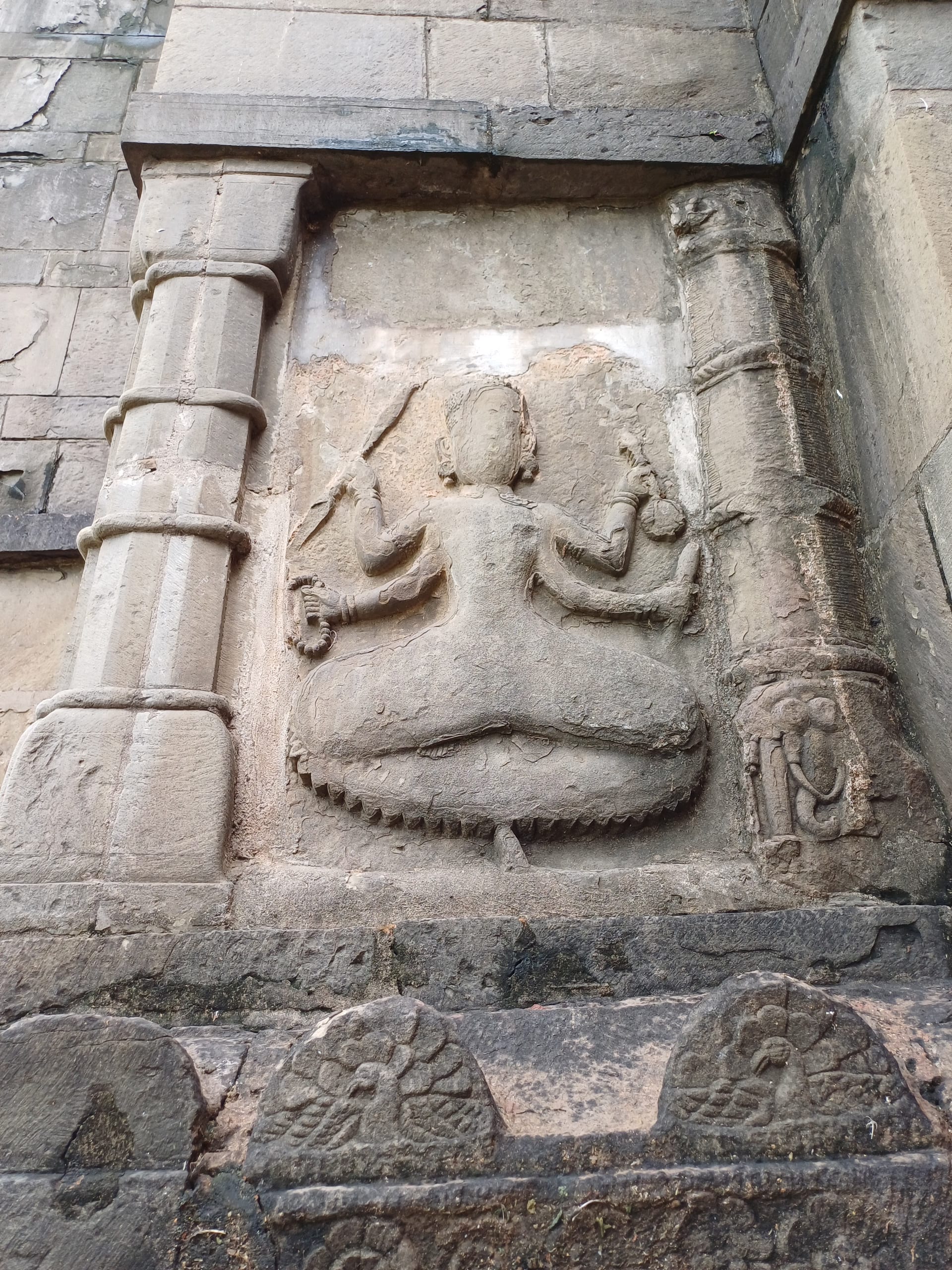Carved stone relief of a seated four-armed deity in a sunken wall niche, raised on a lotus-petalled seat between two engaged columns, with a frieze of small roundels along the base