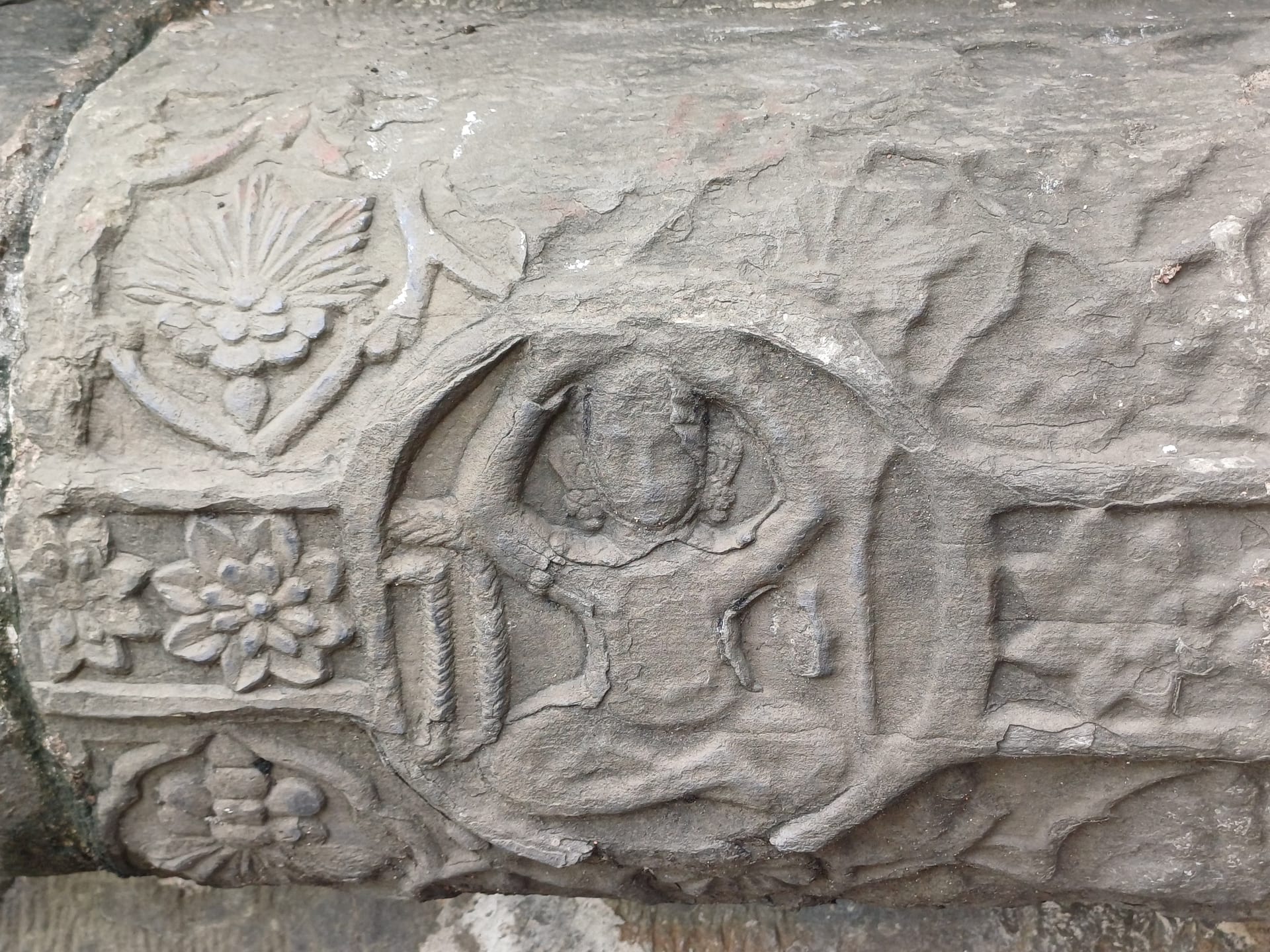 Carved stone detail on a curved temple beam showing a small figure with raised arms inside a circular medallion, beside a lotus rosette and a band of stylised flowers