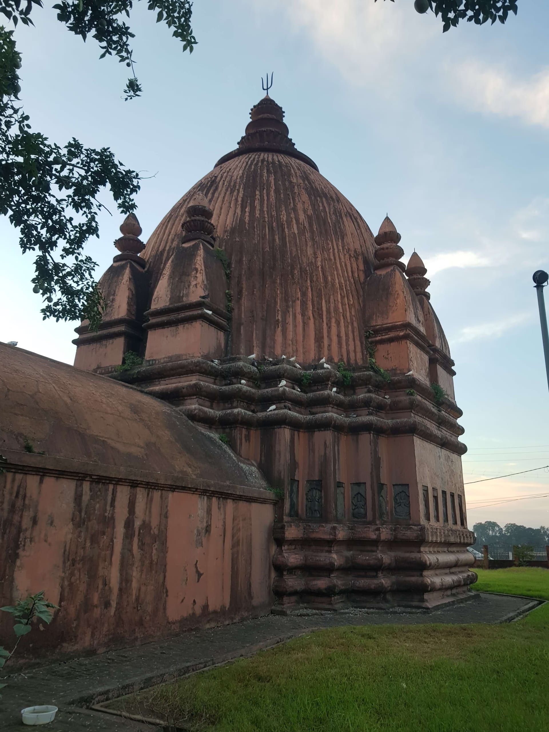 A red-brick Ahom temple seen three-quarter from below, its tall ribbed bell-shaped tower flanked by smaller corner turrets, pigeons perched on the cornices, against a soft pale-blue evening sky
