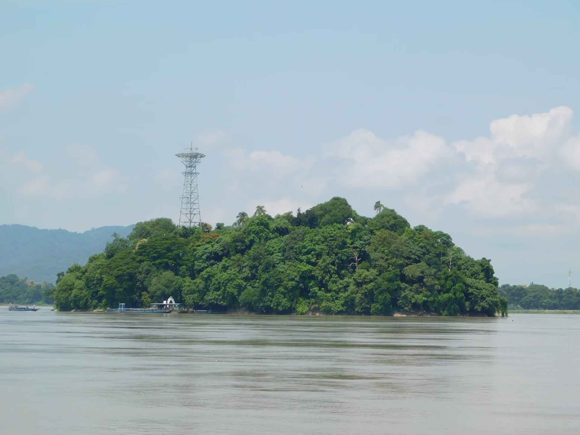The wholly wooded Peacock Island rising from the broad Brahmaputra under a blue sky, the ferry landing at its near shore and low hills along the far bank