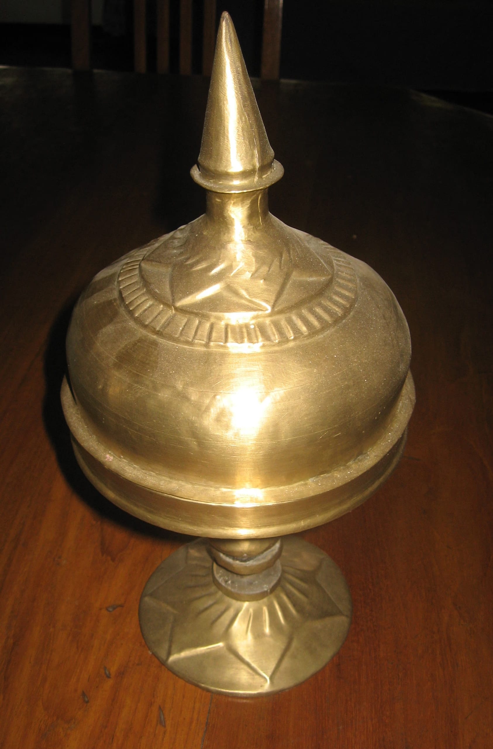 A golden covered bell-metal xorai with a tall conical finial on the lid, the dome and foot engraved with star and ray ornament, standing on a dark wooden surface