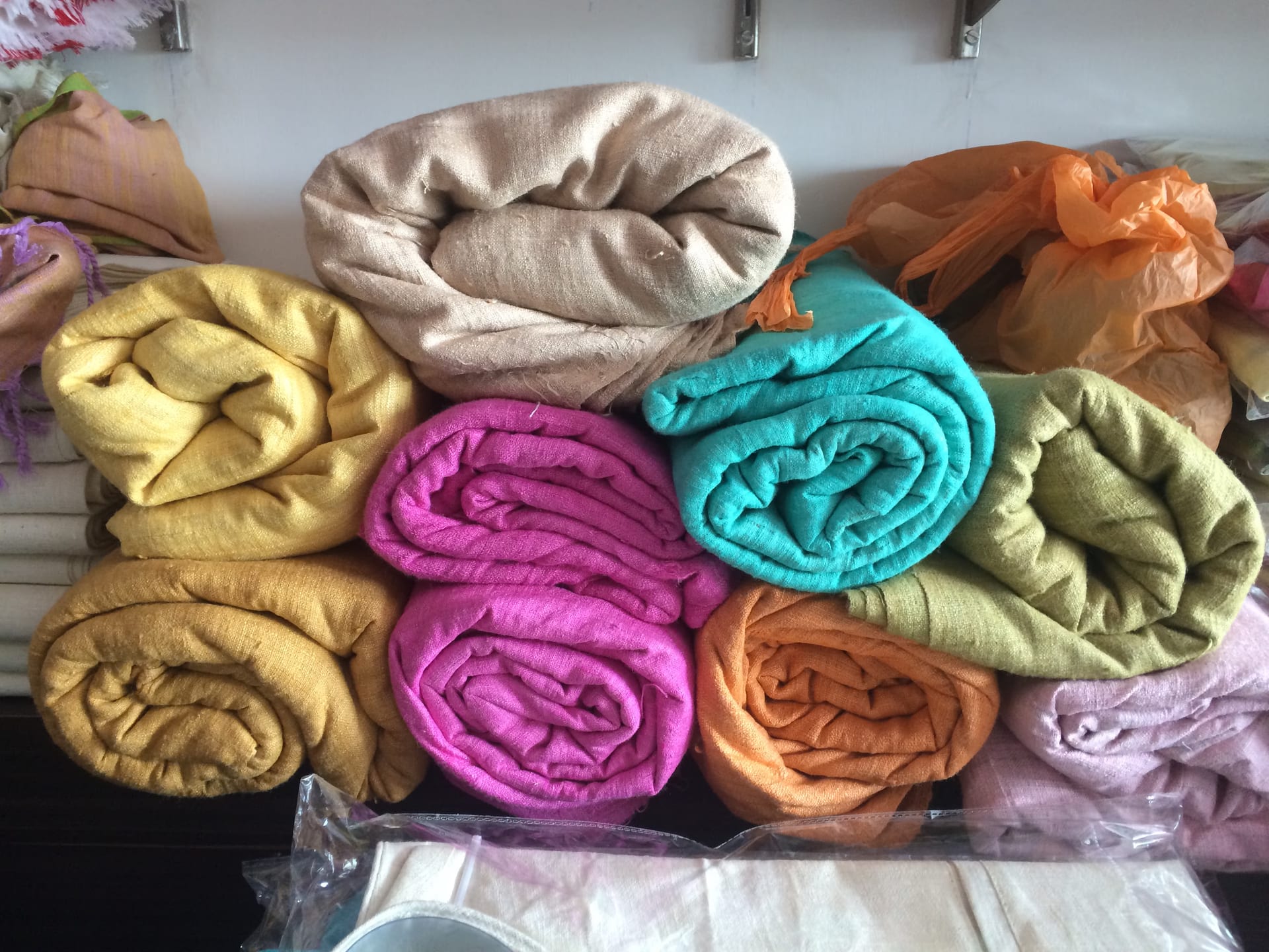 Rolled bolts of eri silk fabric in cream, mustard, pink, teal, ochre and green stacked on a shelf