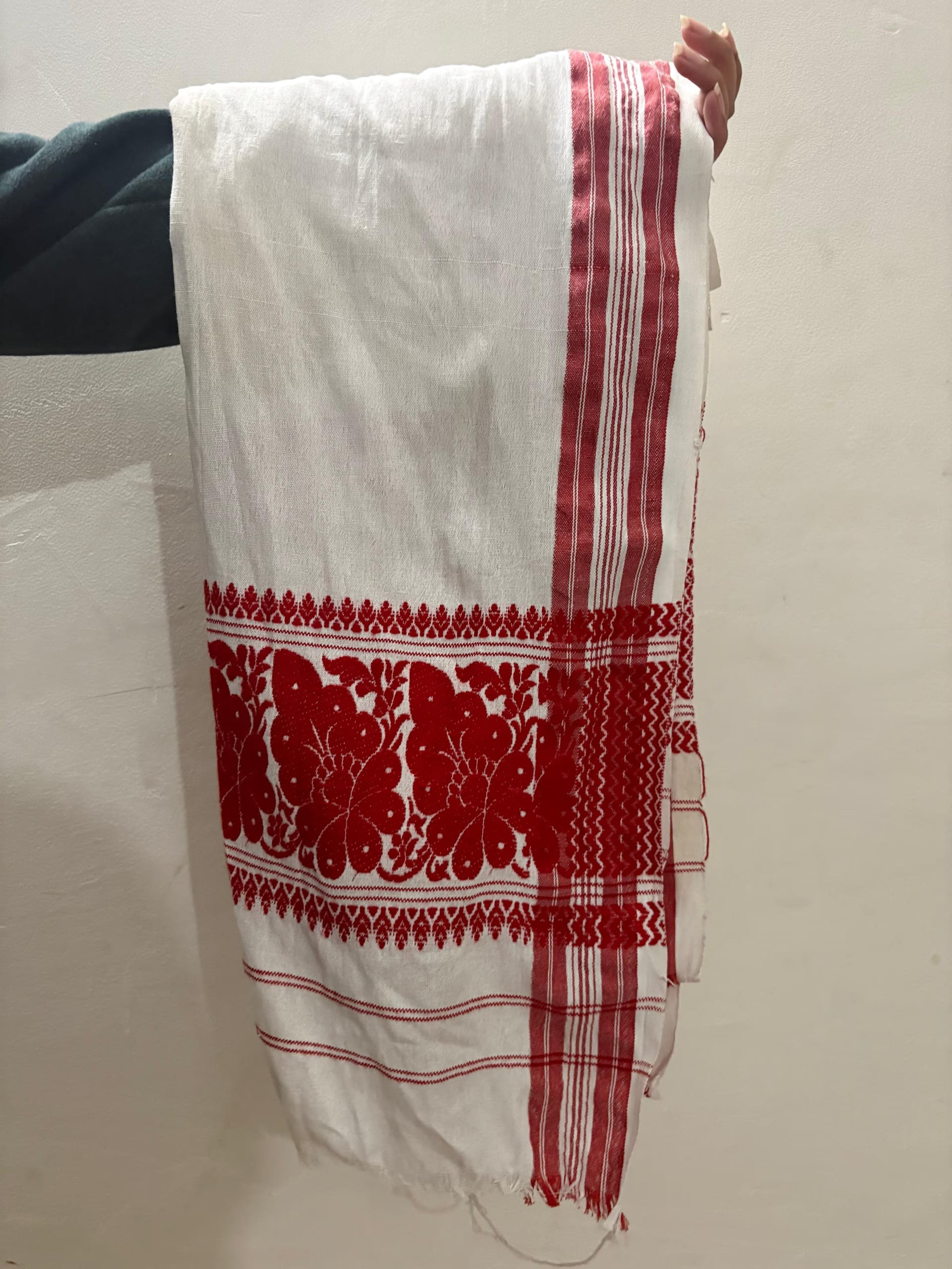 A handwoven white cotton gamosa with red end-borders and a broad band of red woven floral motifs, held up against a pale wall