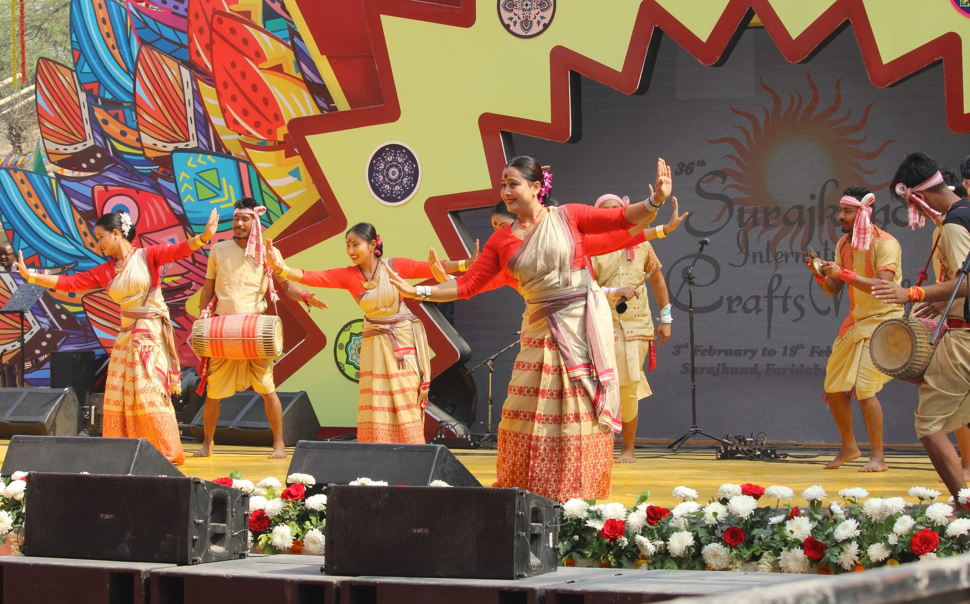 Bihu dancers performing on a stage, the women with a gamosa draped across the torso and the men wearing gamosa headbands while playing the dhol
