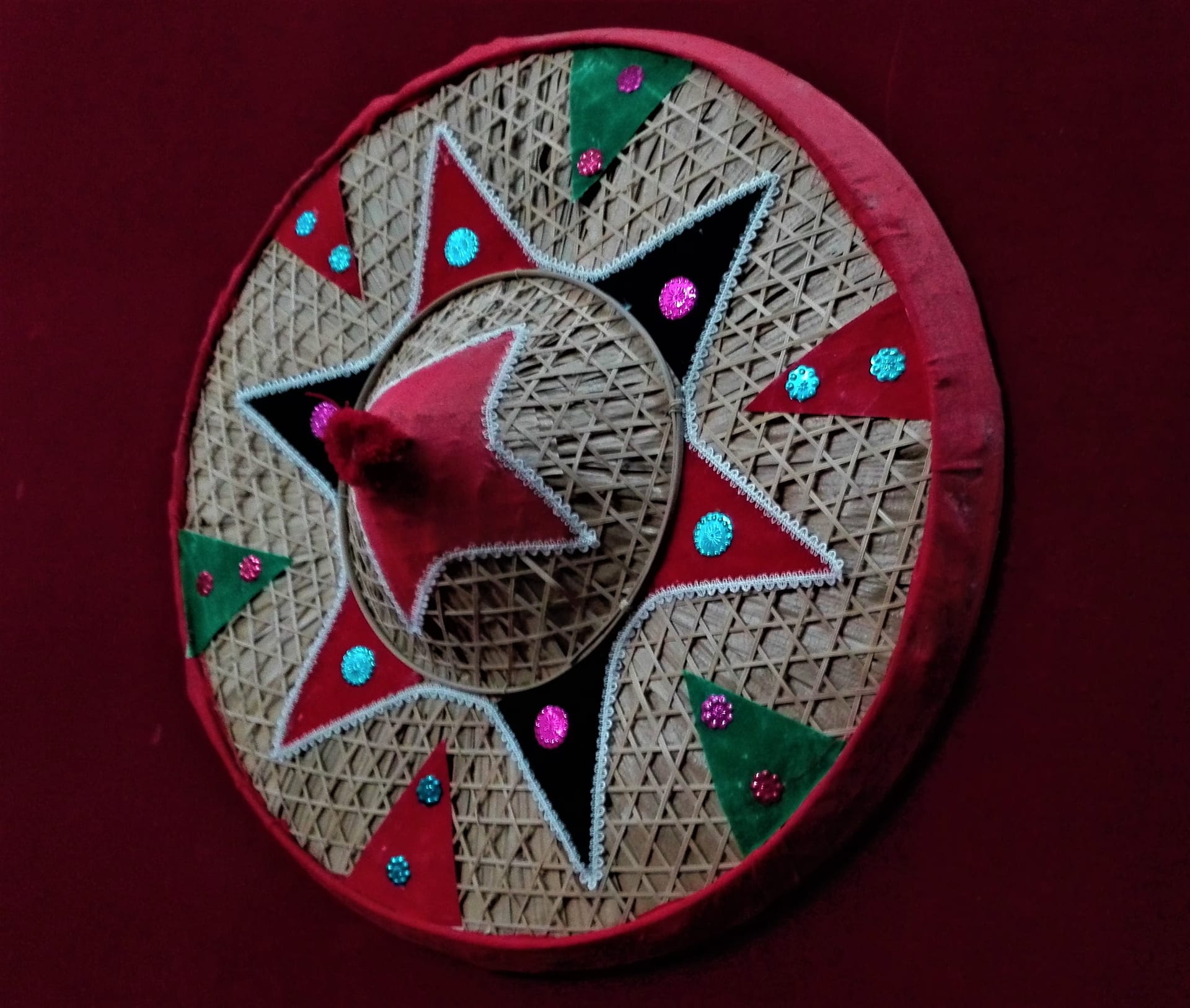 A round phulam japi faced in woven bamboo with a red rim, a green-and-red appliqué star, small mirrors and sequins, set against a deep maroon backdrop