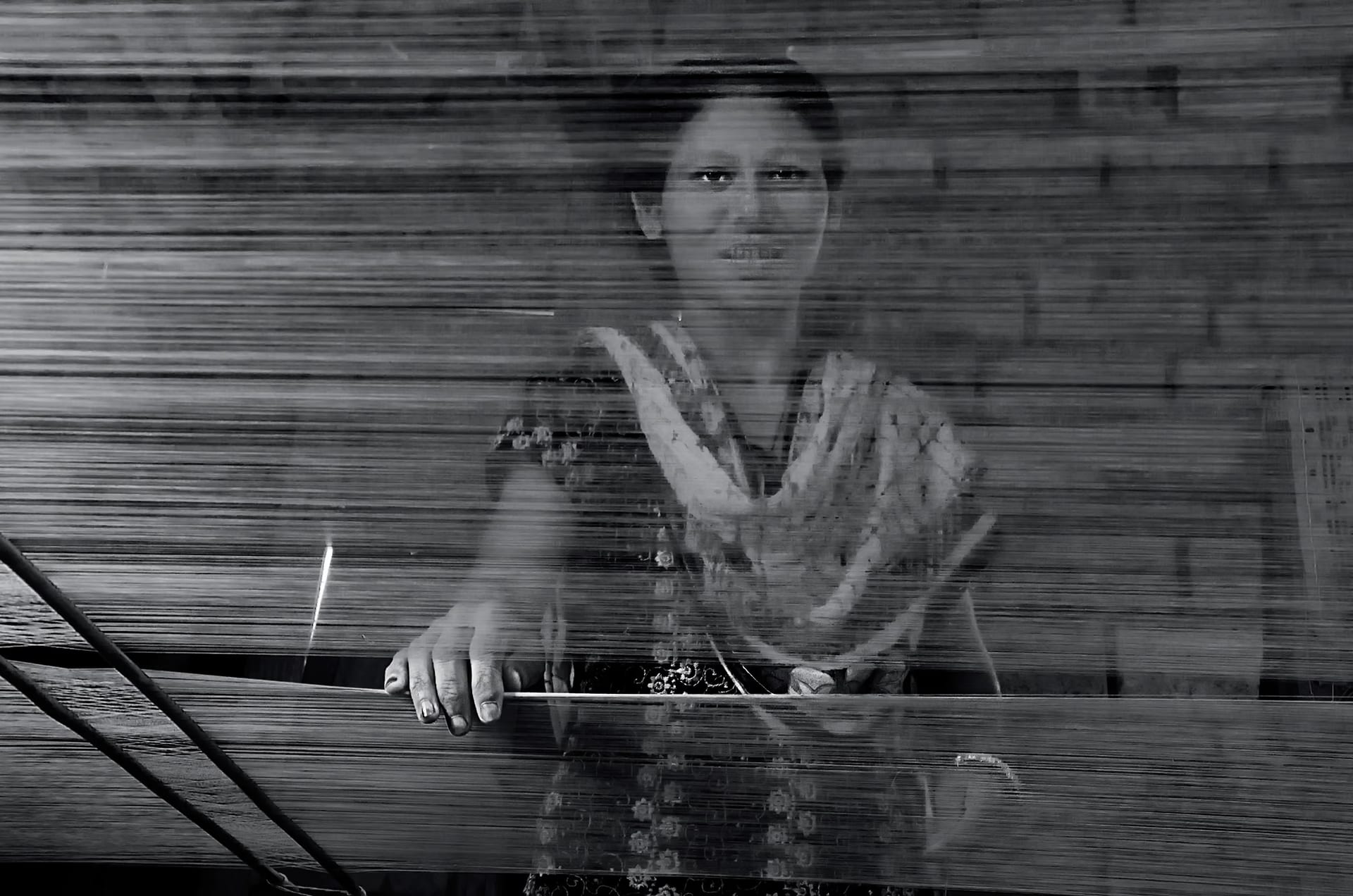 A weaver seen through the stretched warp threads of a handloom, her hand resting on the loom beam, in Sualkuchi