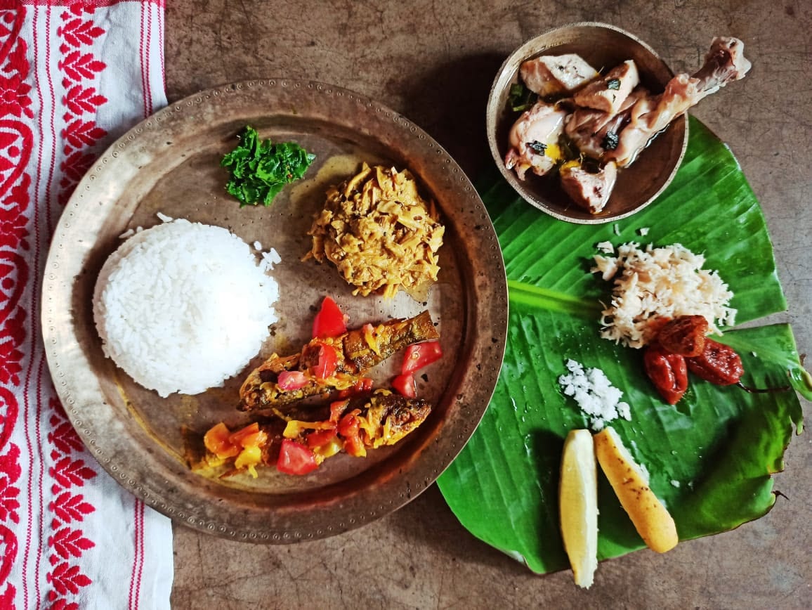 An Assamese lunch on a bell-metal plate with rice, fish, and condiments, beside a banana leaf