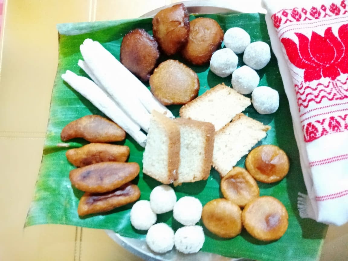 A banana-leaf platter of til pitha, fried tel pitha, golden ghila pitha and white coconut laru beside a red-and-white gamosa
