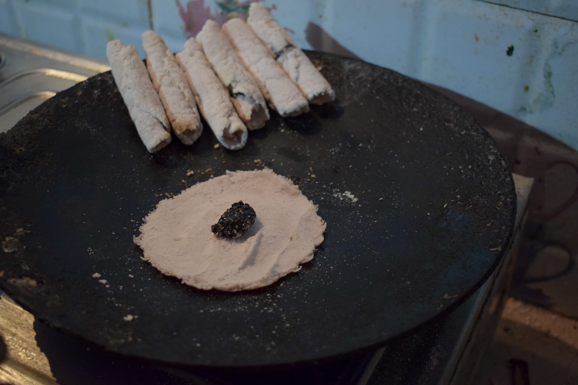 Rolled til pitha at the back of a hot griddle and, in front, an open round of rice-flour batter with a dollop of dark sesame-and-jaggery filling ready to be folded