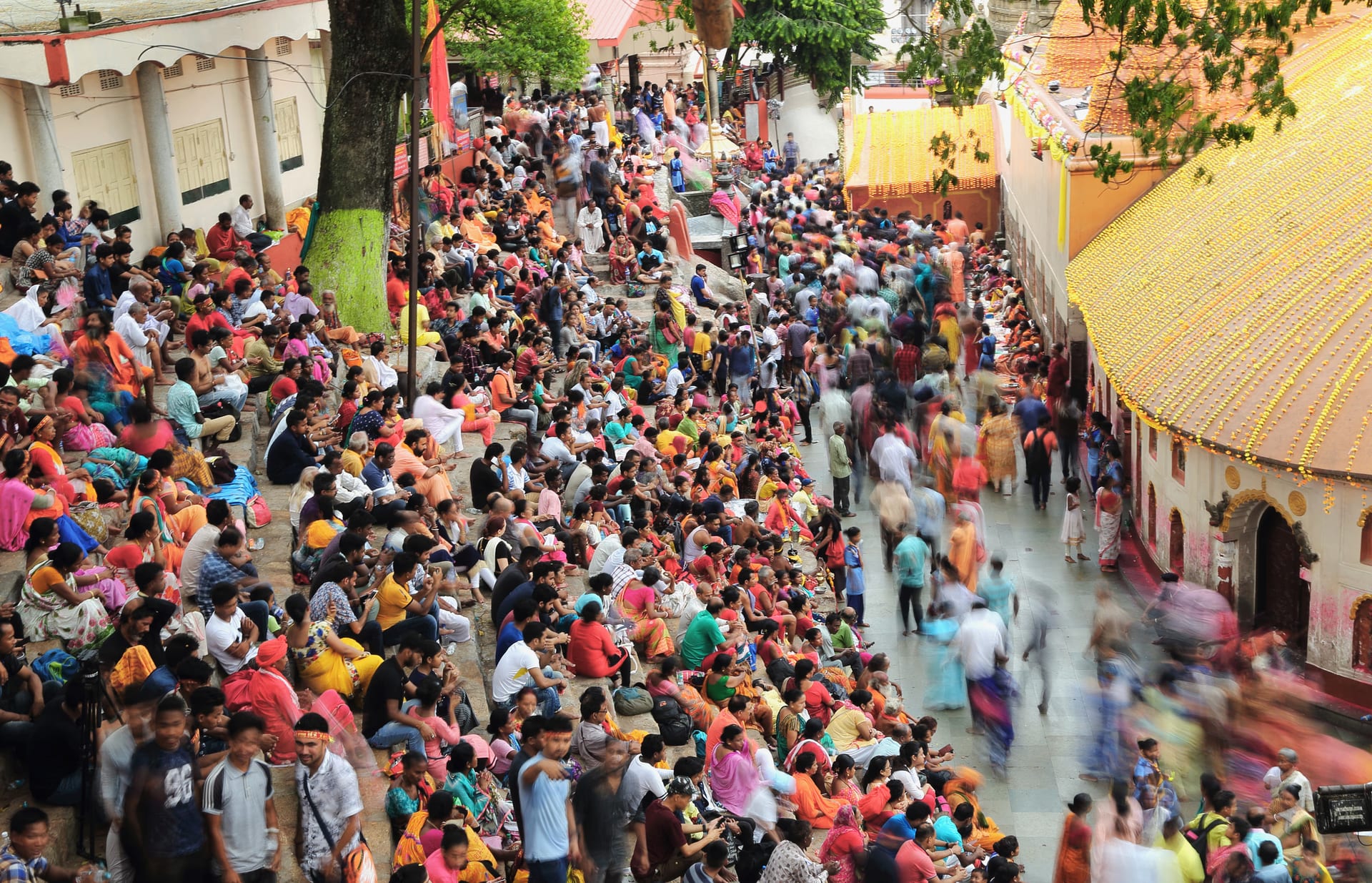 Thousands of pilgrims in bright clothes packed on the stone terraces beside the Kamakhya temple, its curved roof hung with marigold garlands, walkers blurred in motion below
