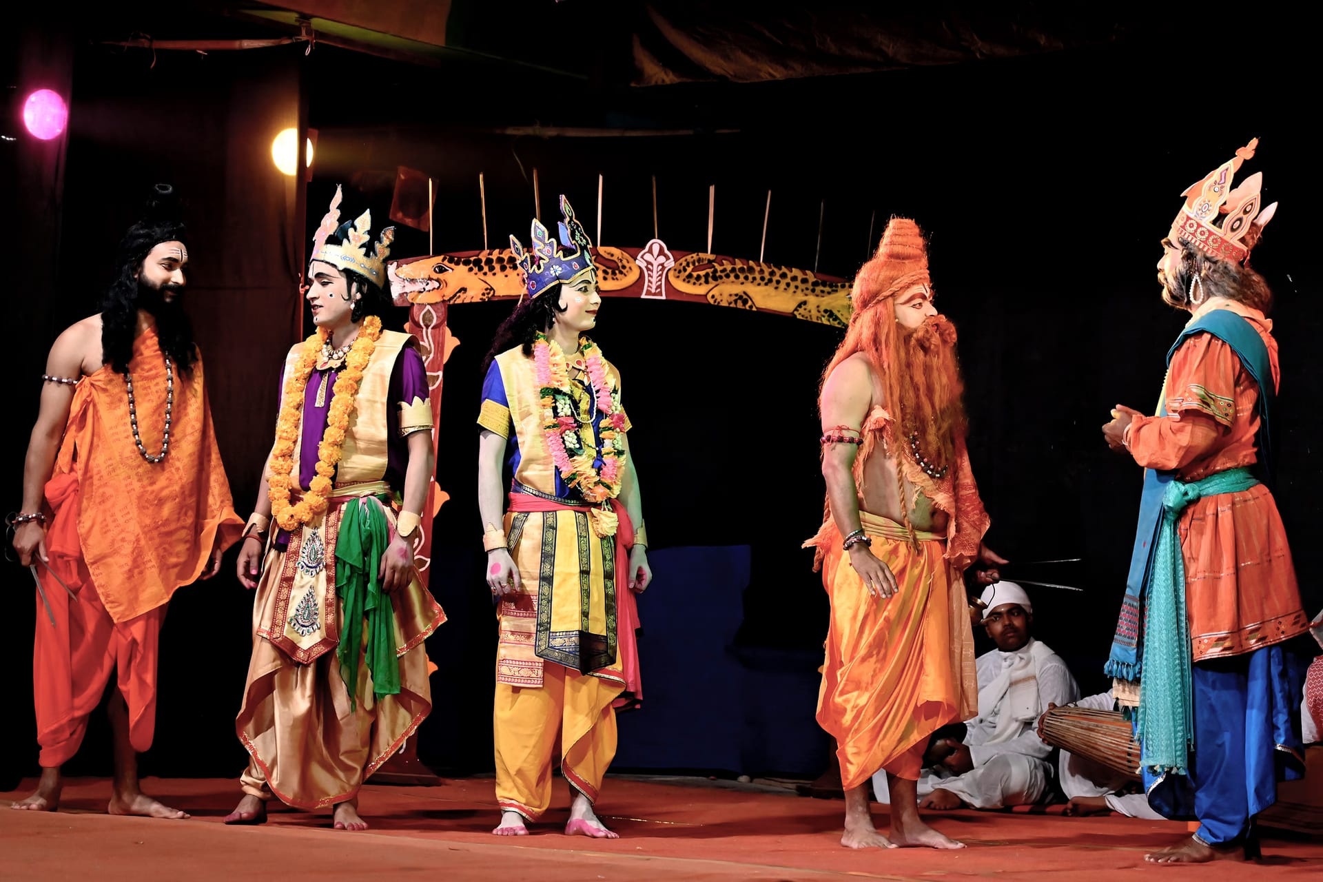 Costumed performers in robes, garlands and crowns stand on a sattra stage with a khol drummer seated behind them