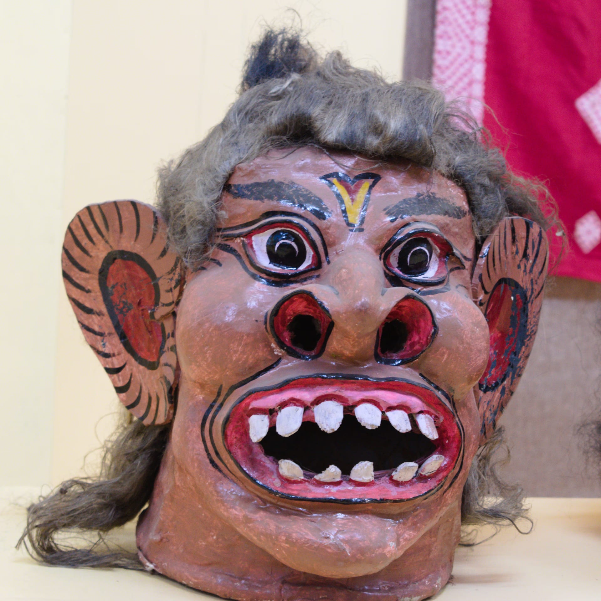 A painted papier-mache and clay mukha face-mask of a fierce demon, with a brown face, bulging red-rimmed eyes, a gaping red mouth showing white teeth, large painted side-ears and grey hair, displayed against a plain pale museum wall