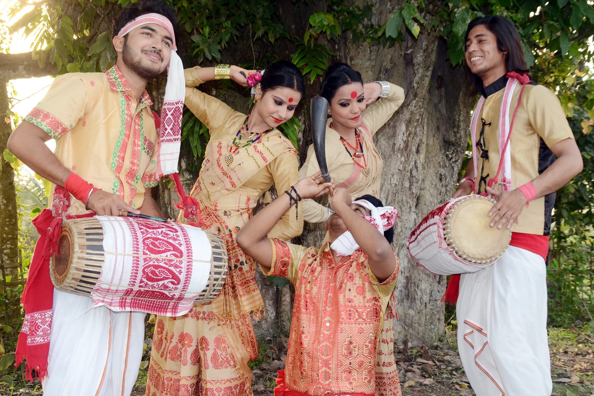 A posed group under a tree: two young men in cream kurtas and red-bordered gamosa each carrying a dhol slung at the waist, flanking two women in cream-and-red Bihu dress with hands raised to their hair, while a third man kneeling in front blows a buffalo-horn pepa