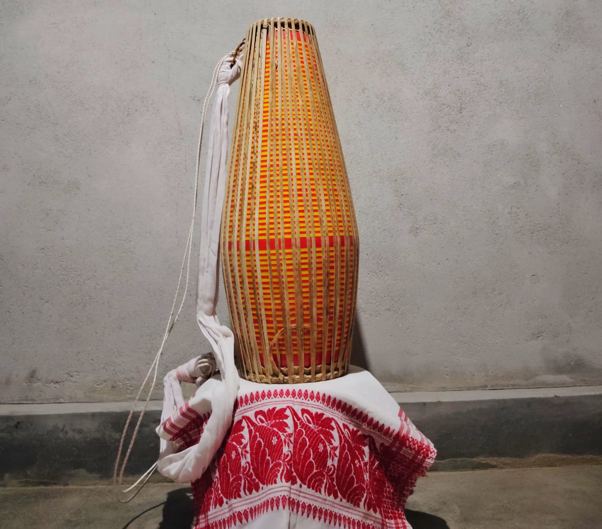 A single two-headed khol drum standing upright on its narrower head, its long tapering body wrapped in close vertical bamboo-and-cane lacing over orange and red banding, a white cloth carrying strap looped down one side, the drum resting on a folded red-and-white gamosa against a plain grey wall