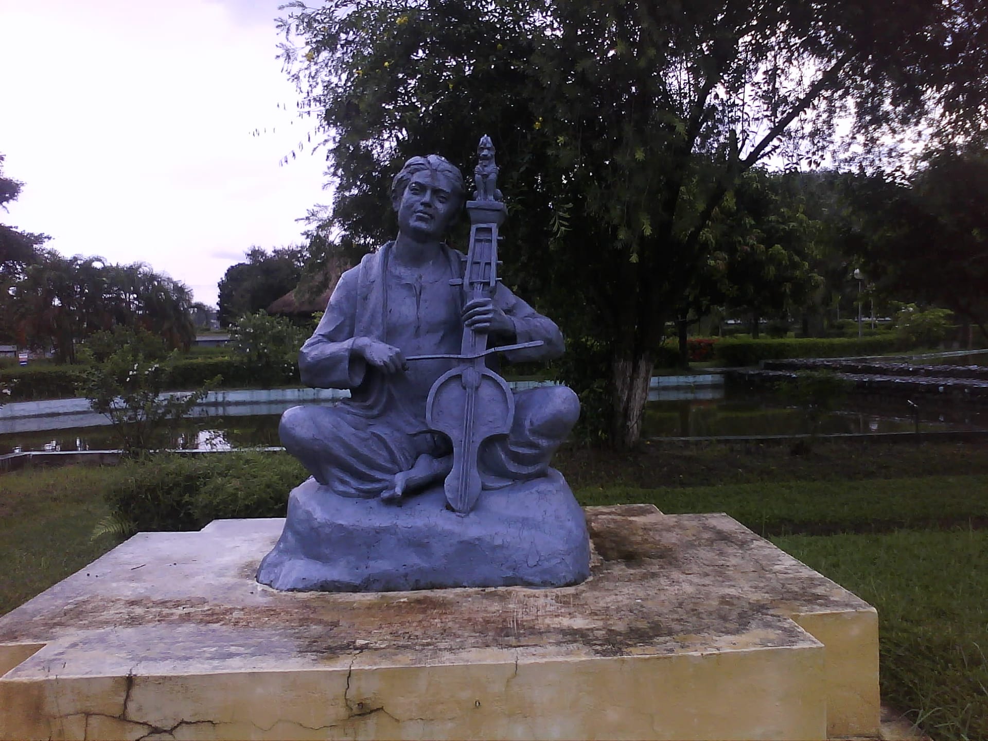 A grey stone seated statue of Sankardev cross-legged on a plinth, playing a long-necked stringed instrument held upright, with trees and a pond behind