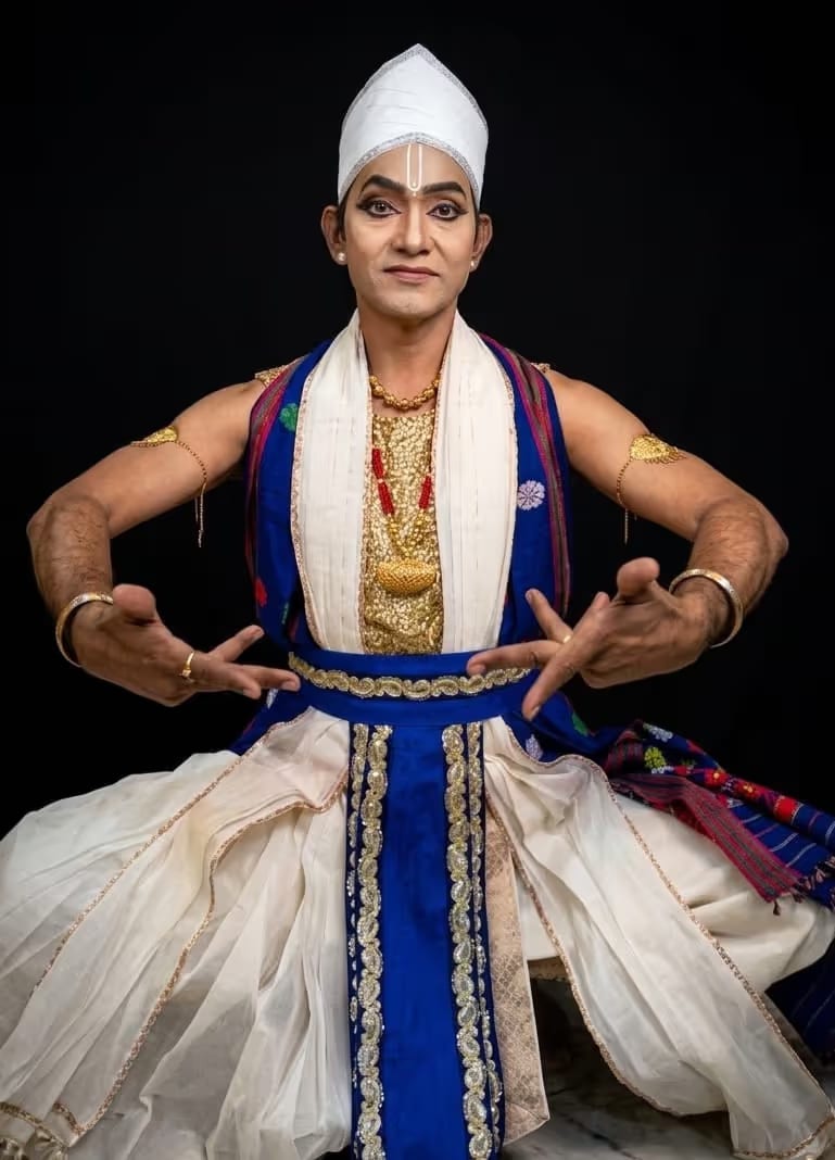 A male Sattriya dancer kneeling in a white dhoti and chadar with a blue and gold waist-wrap and pleated skirt, a folded white paguri turban and gold ornaments, hands held in a mudra against a dark background