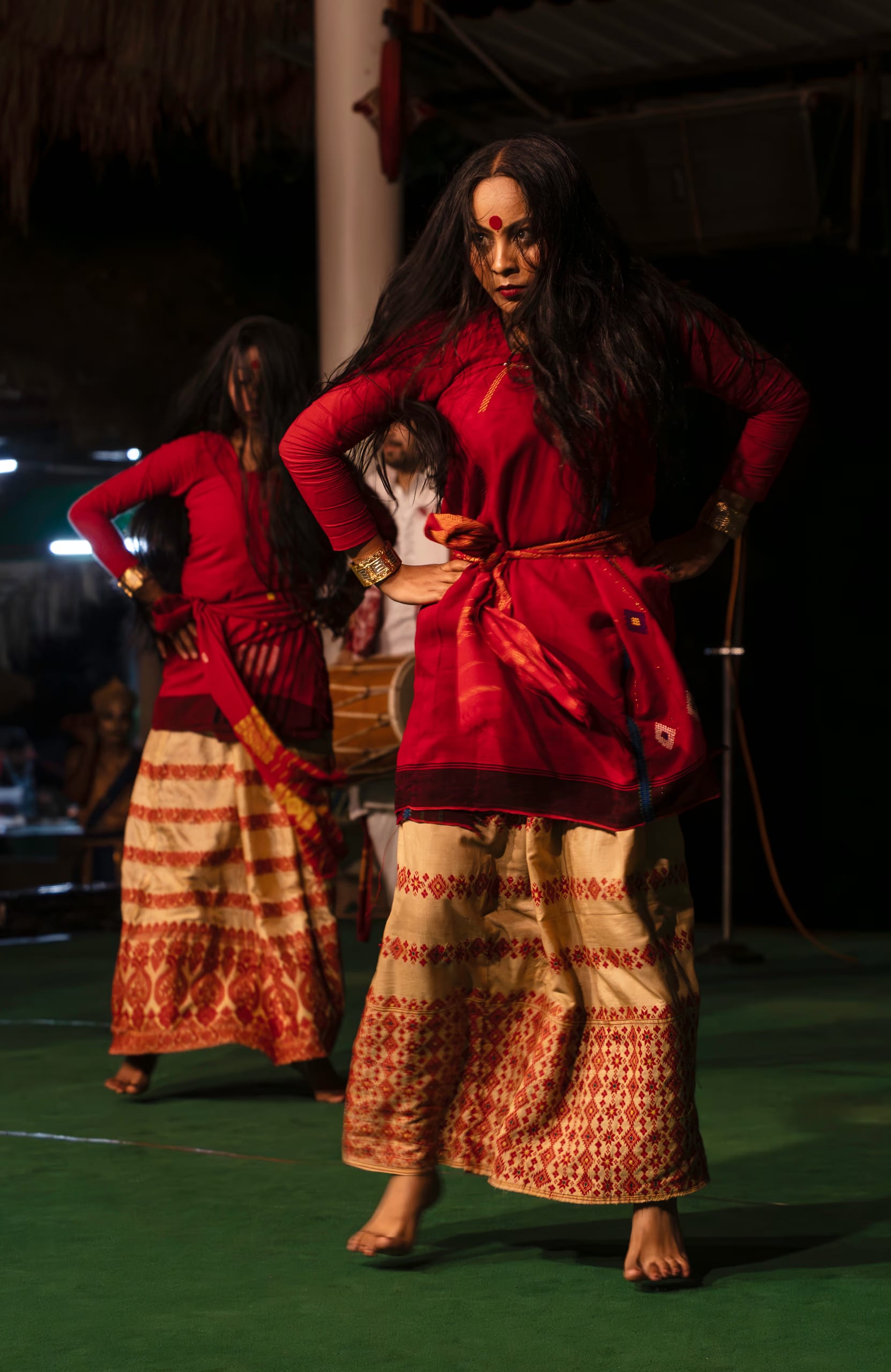 A Sattriya dancer in a red blouse and cream Muga silk skirt woven with traditional Assamese motifs, gold cuff bracelets, a khol drum visible behind