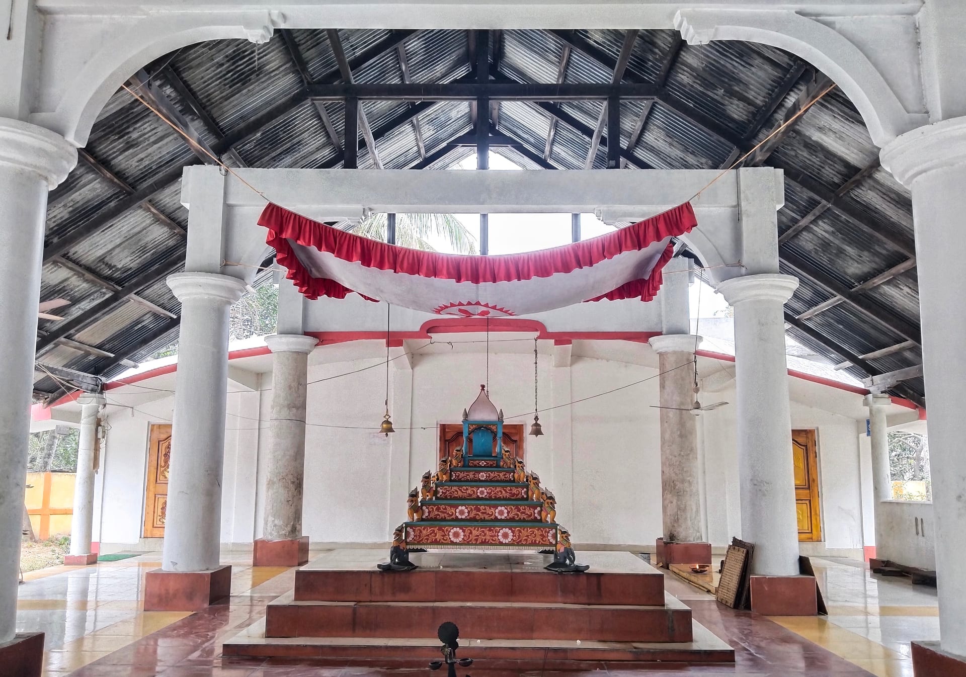 Interior of a Vaishnavite namghar prayer hall with white pillars and a central decorated altar throne flanked by hanging bells