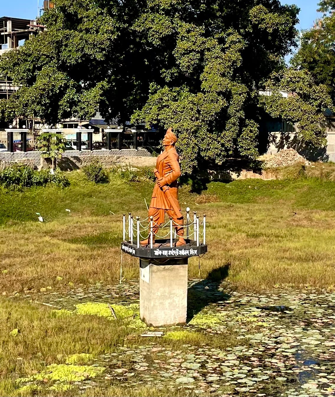 Bronze-coloured statue of a robed standing figure on a plinth set in a tank, with trees behind