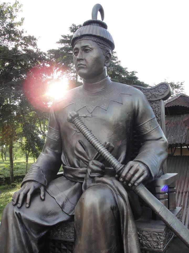 Dark bronze seated statue of Sukaphaa holding a sheathed sword across his lap, in a Tai tunic and ridged conical crown, with tea-garden trees behind