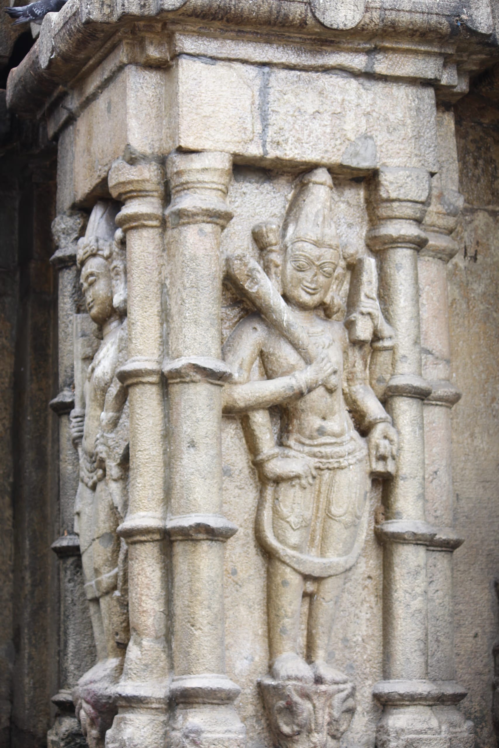 A weathered grey stone relief of a standing crowned deity figure carved on the exterior wall of the Kamakhya temple, set between two slender carved pilasters, one arm raised