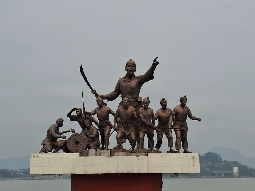 Bronze statue group: Lachit Borphukan standing tall with a raised sword and an outflung pointing arm, a band of Ahom warriors and a seated gunner with a cannon arrayed below on a white plinth, the Brahmaputra and far hills behind