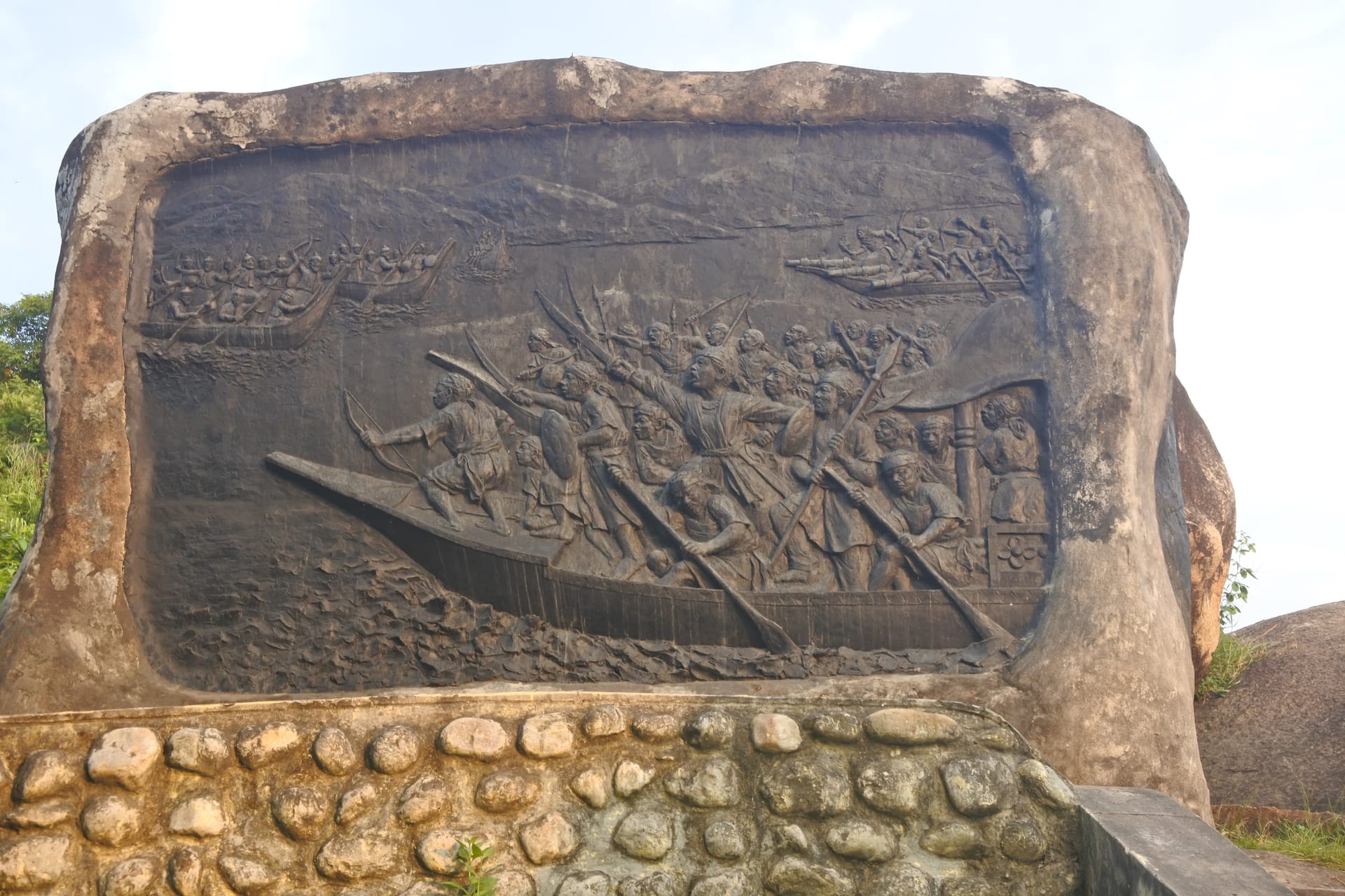 Rock-carved relief of Ahom war-boats packed with spearmen and rowers under a pointing commander, cut into a large grey boulder