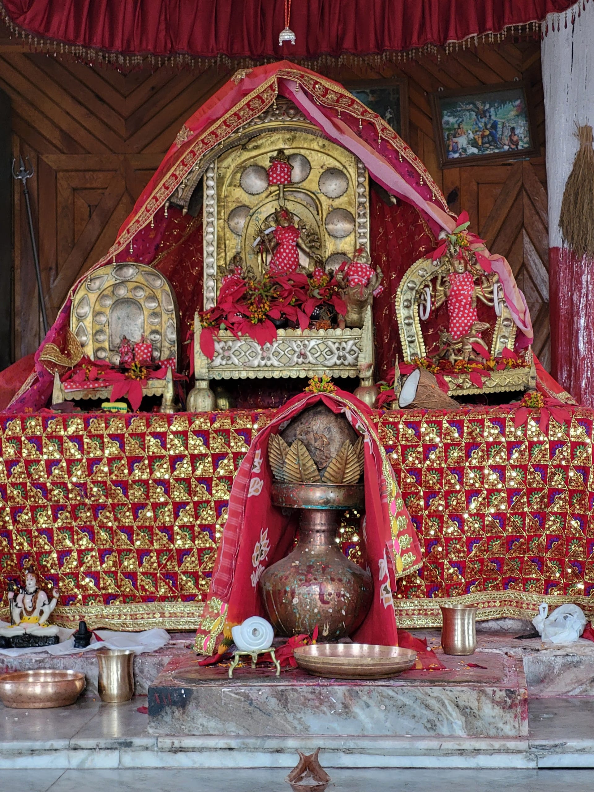 Temple sanctum draped in red and gold-sequined cloth: three gilded thrones bearing deity figures behind a central decked sheaf-like form, with brass lamps, a copper kalash and a copper offering plate set on a marble platform in front.