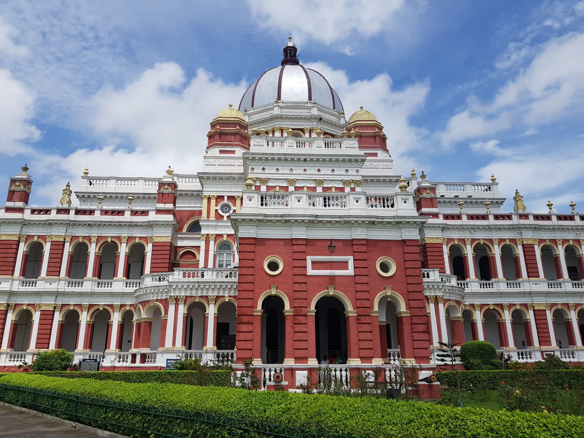 Frontal view of the Cooch Behar Palace, a red-brick and white classical building with a central metal dome over an arcaded façade