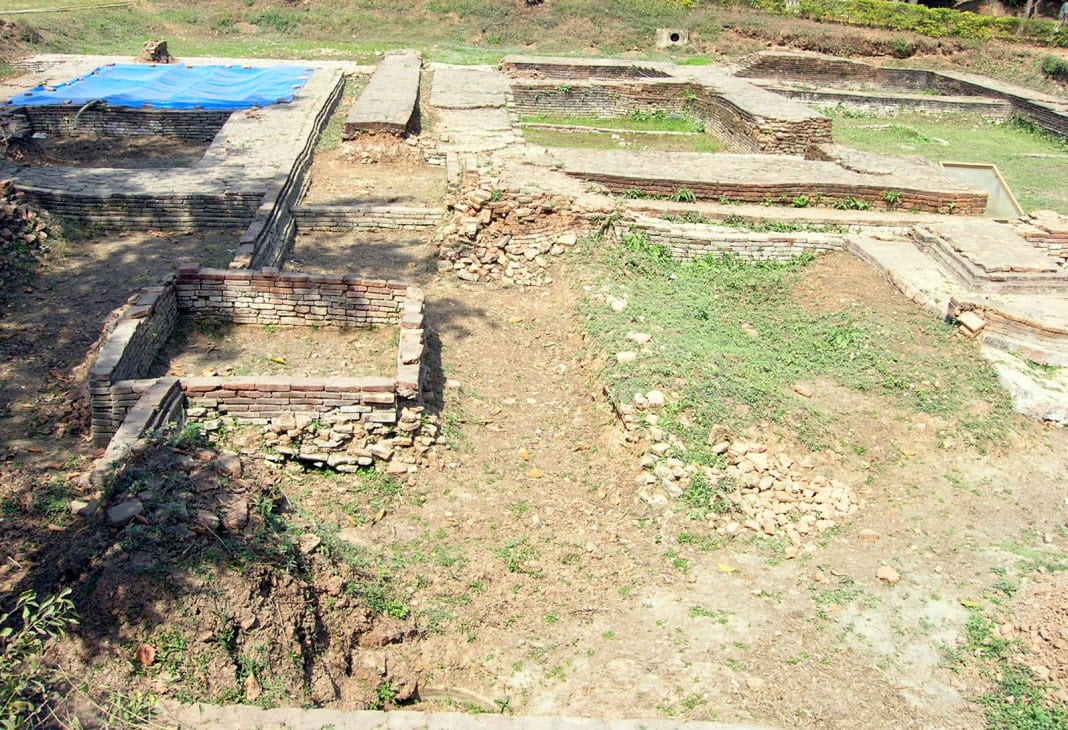 Excavated brick foundations at the Ambari site: low walls of laid brick, a stepped tank structure to the right, cleared earth between them, part of the dig under a blue tarpaulin