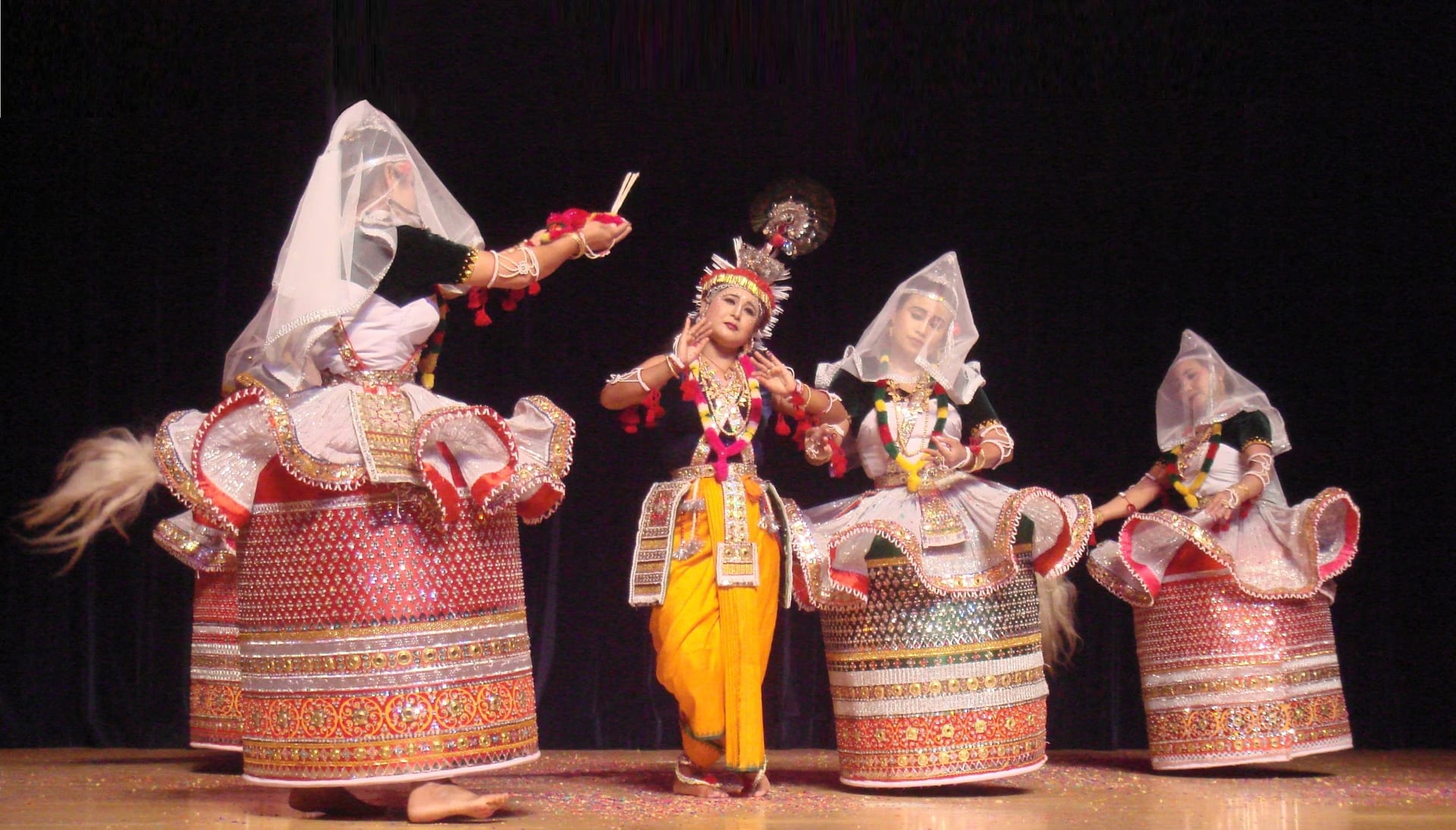 Manipuri Ras Lila dancers: a central figure costumed as Krishna with a flute, flanked by three gopis in the stiff, mirror-worked cylindrical Kumil skirt and translucent white veil