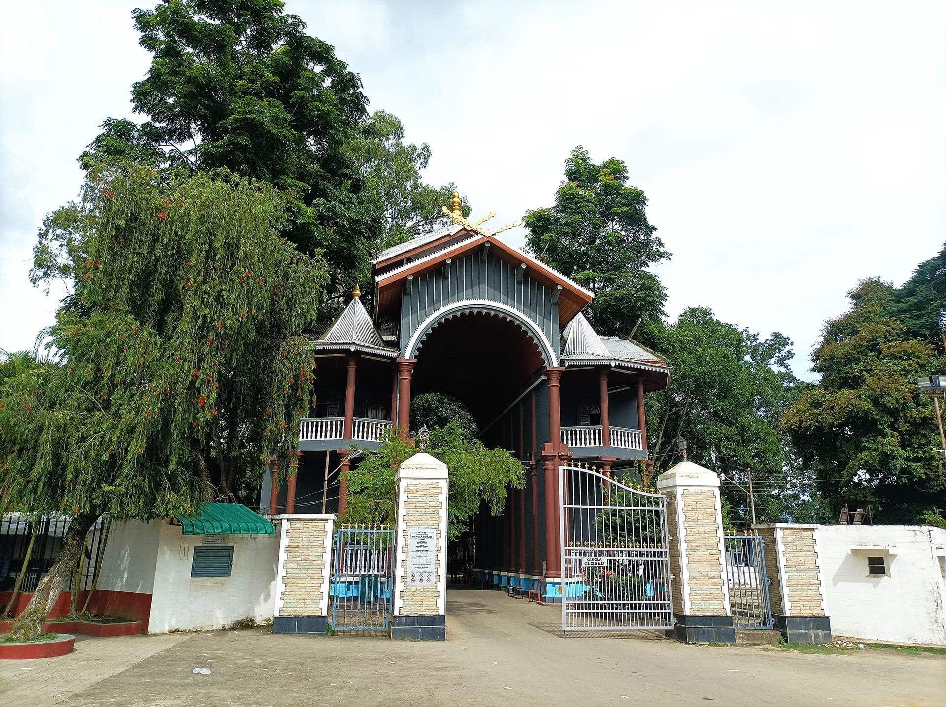 An arched ceremonial gate-pavilion with twin turreted balconies and a steep roof, set behind a fenced compound