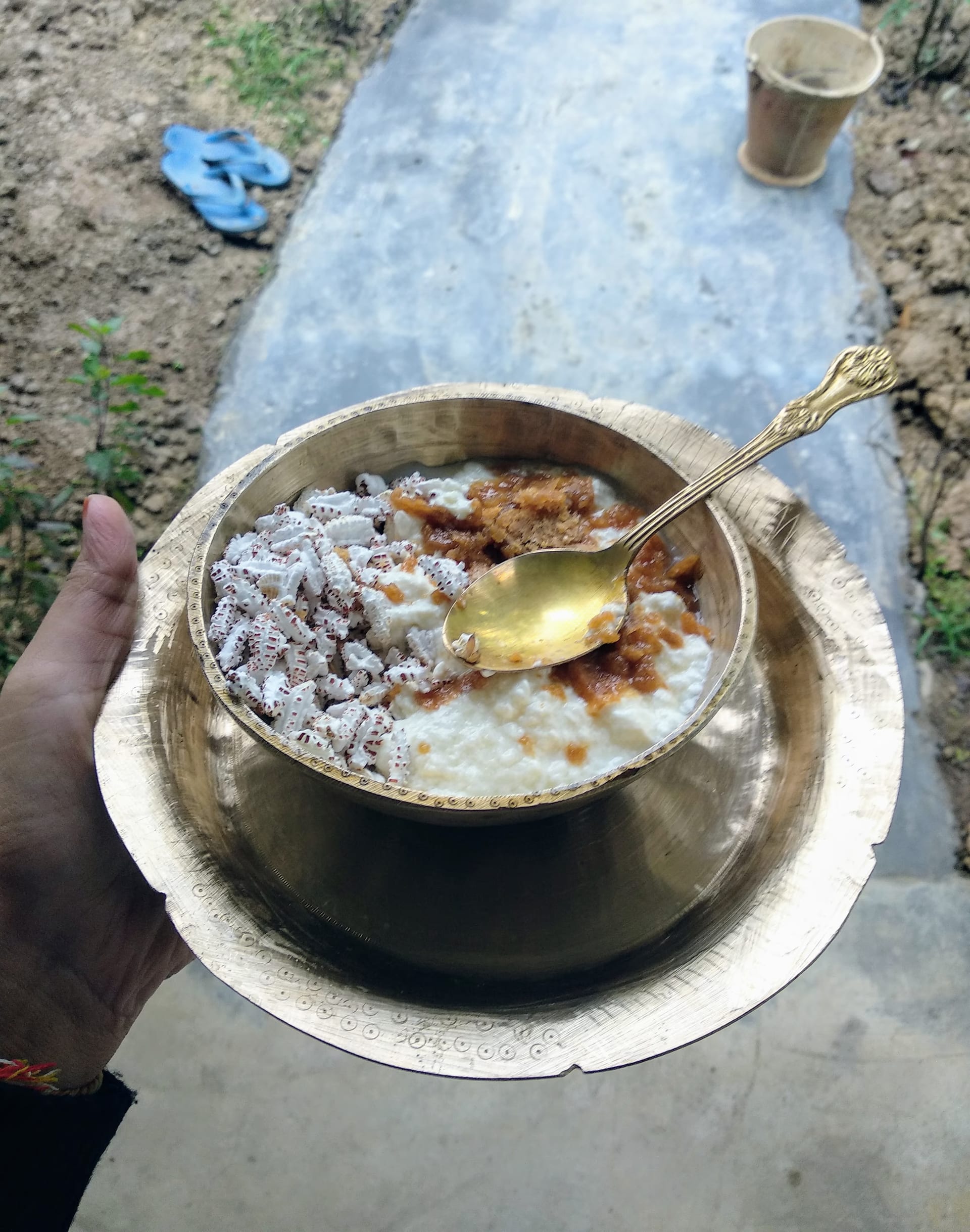 A hand holds a bell-metal bowl of curd mixed with puffed rice and jaggery, a spoon resting in it