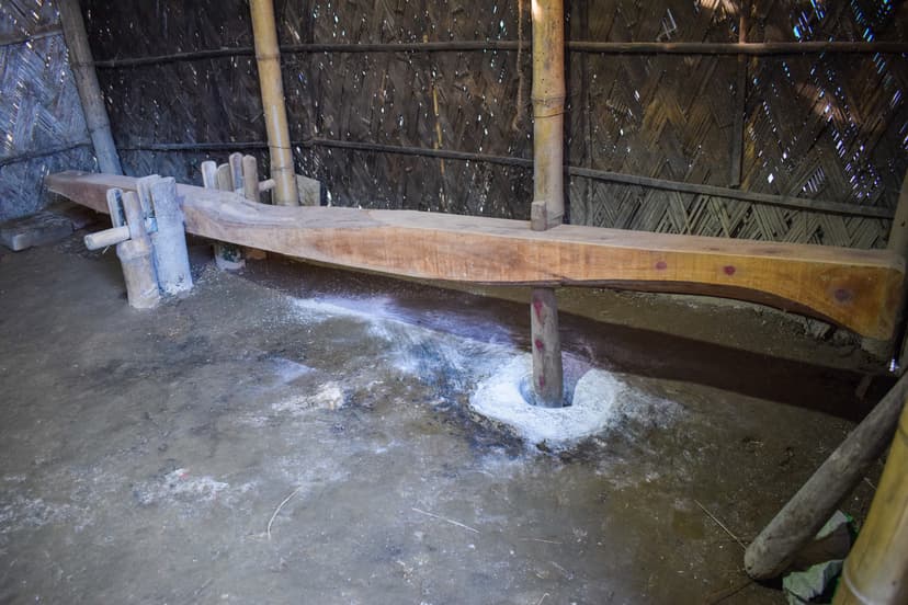 A wooden dheki, a foot-operated lever pestle, set over a floor mortar inside a bamboo-walled house