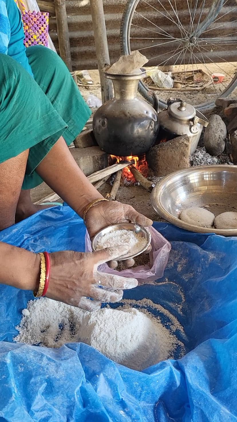 A vendor's hands scoop rice flour beside a brass tekeli pot steaming over a wood fire, with finished round cakes on a tray