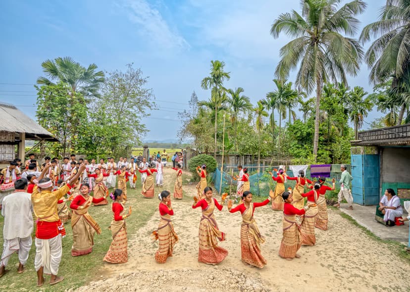 Young women in golden muga mekhela sador and red blouses dancing in a ring in a village yard, with male drummers at the edge