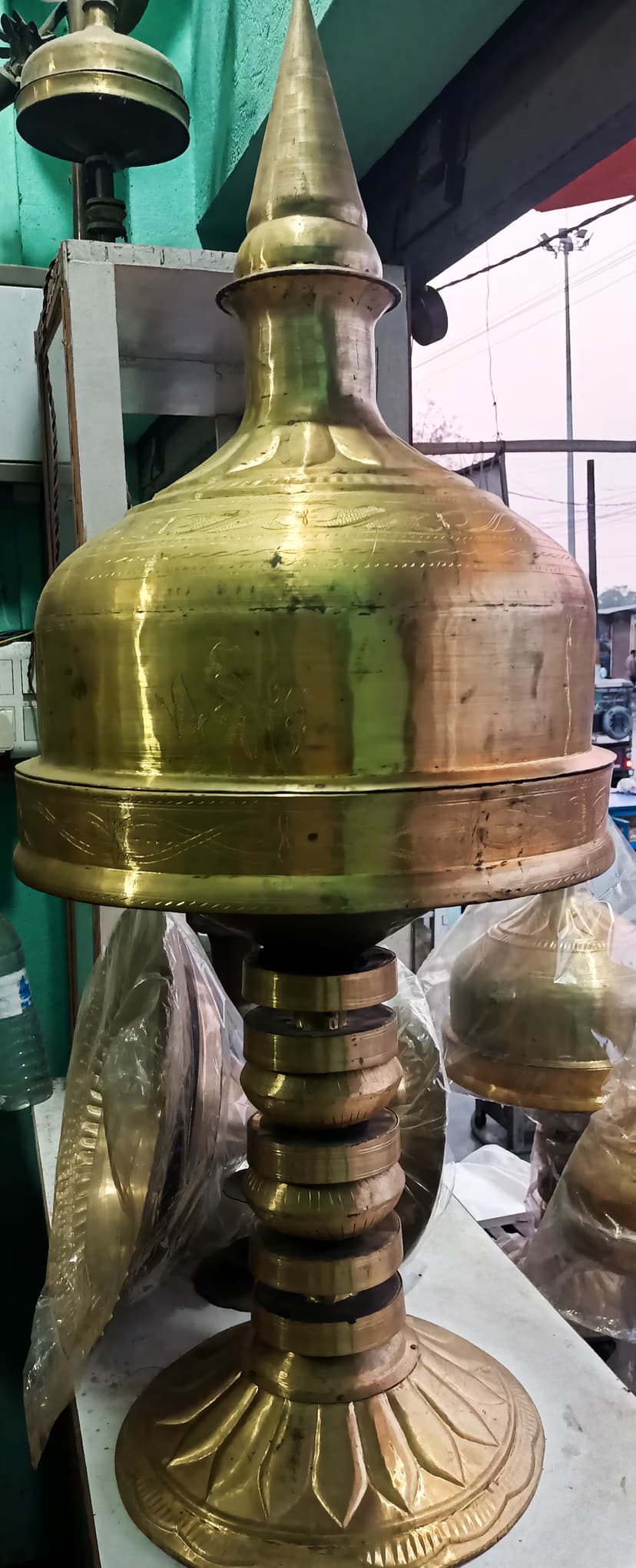 A tall polished brass xorai with a domed lid and lotus-petal base, set on a white workbench with wrapped metalware behind it
