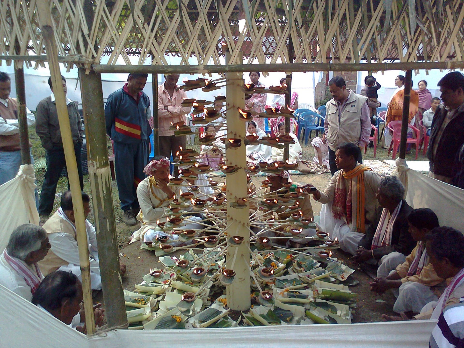 Under a bamboo pavilion, Tai-Ahom men sit around a tall stem bristling with lit oil lamps on leaf trays, with elders praying in the foreground and a crowd behind