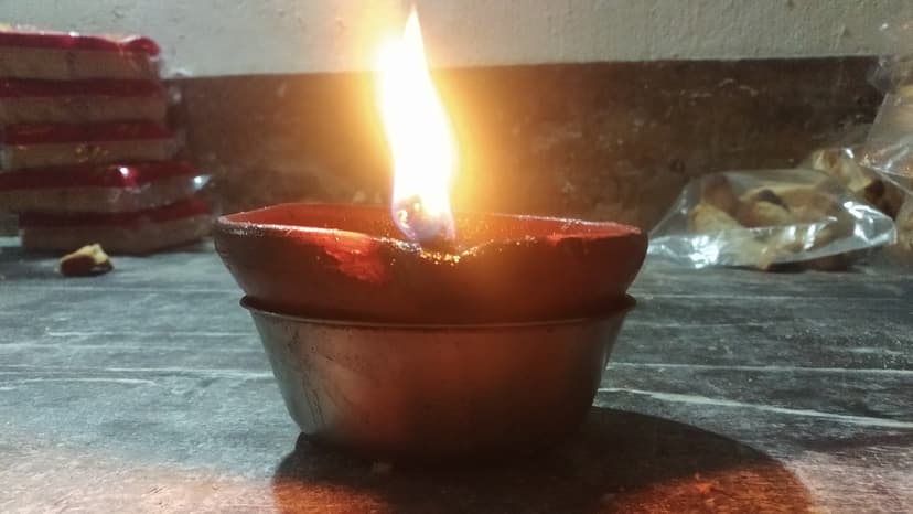 A single lit earthen oil lamp with a tall bright flame on a dark stone floor, glowing warmly against a shadowed background