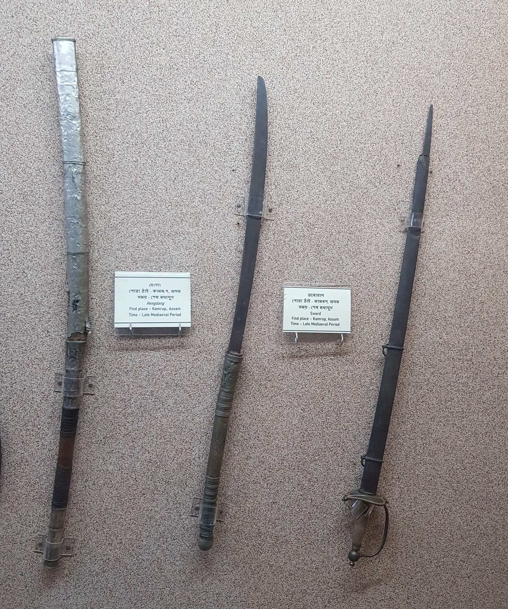 Museum wall display of three long Assamese blades: at left a sheathed hengdang in a metal scabbard, in the middle an unsheathed single-edged blade with a ribbed grip, and at right a basket-hilted sword, each beside a printed bilingual label.