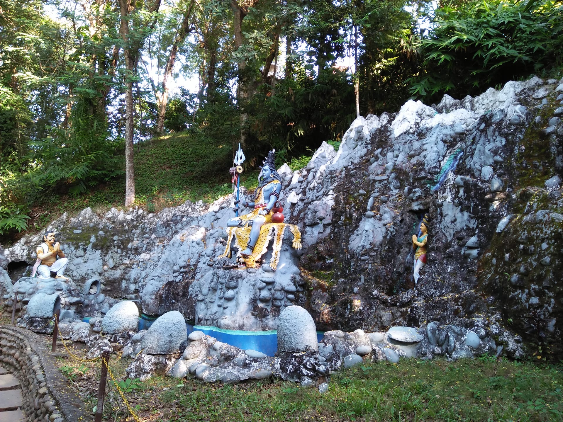 A painted sculpture group on a rocky slope: a blue Shiva seated on a tiger skin holding a trident, with a kneeling crowned figure at left and a standing female figure at right, both with hands folded