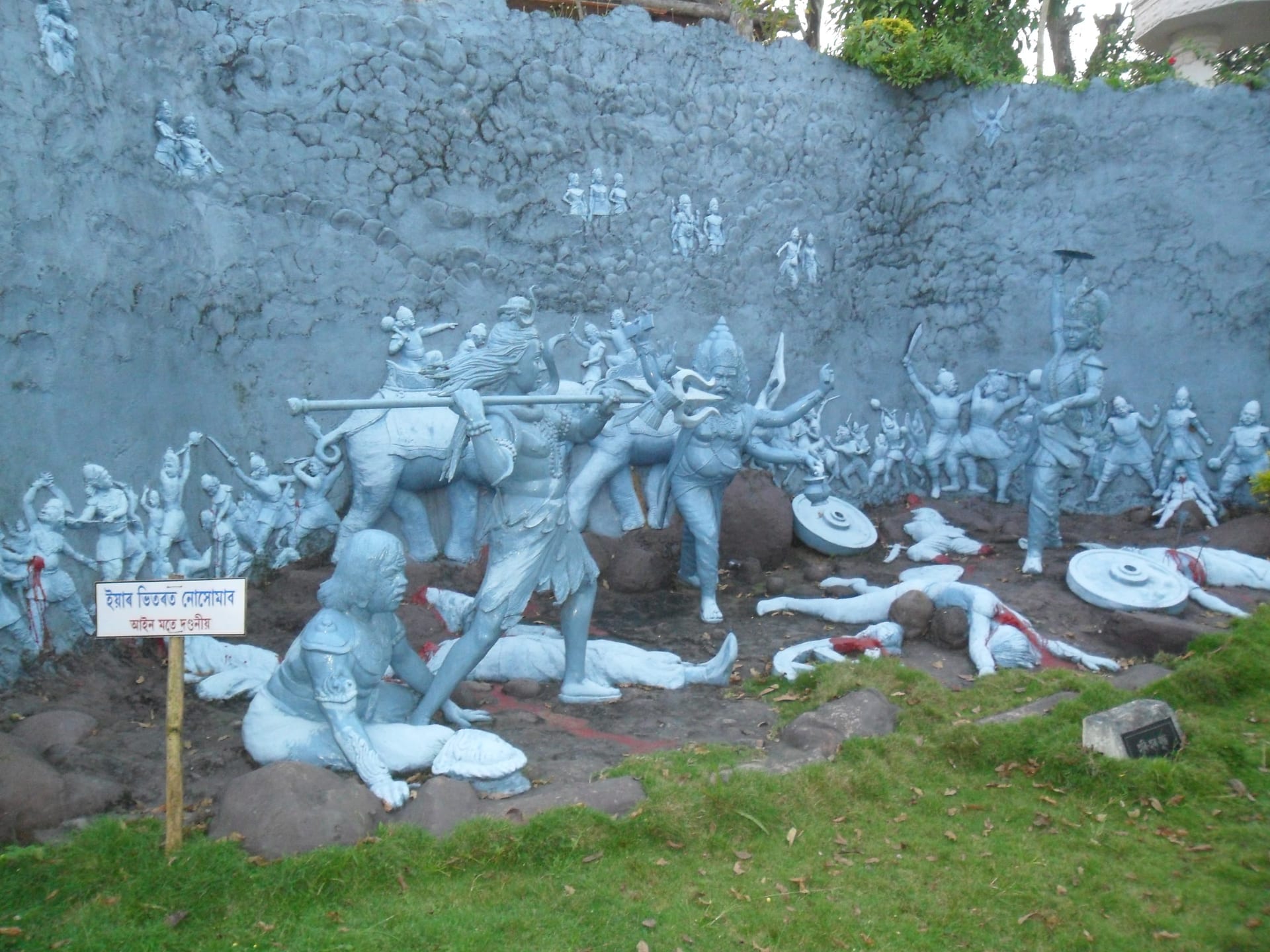 A large grey sculptural tableau against a rock wall: Shiva with raised trident and Krishna on his mount above ranks of fighting figures, with fallen warriors and shields strewn on the ground below, red paint marking their wounds
