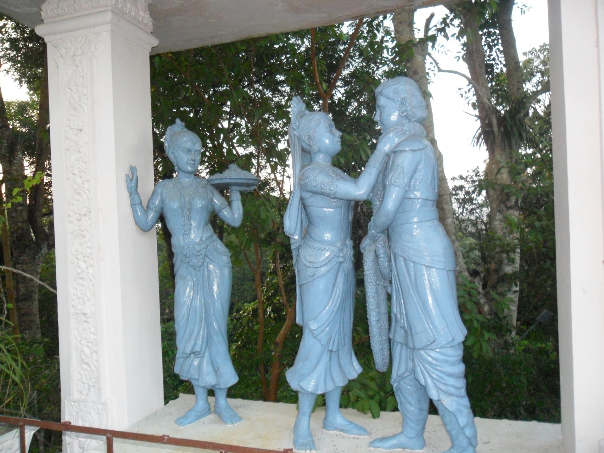 Three pale blue sculpted figures under a white pavilion: a woman holding a tray at left, and at right a woman raising a garland toward a standing man who holds one himself