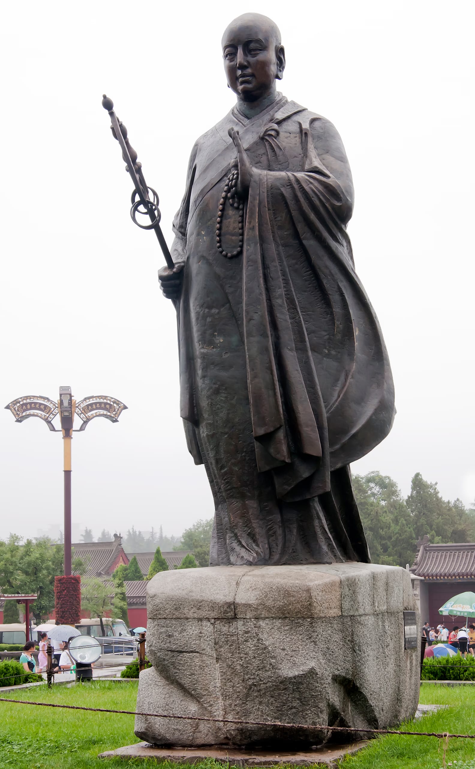 A bronze standing statue of the monk Xuanzang holding a ringed monk's staff and prayer beads on a stone plinth