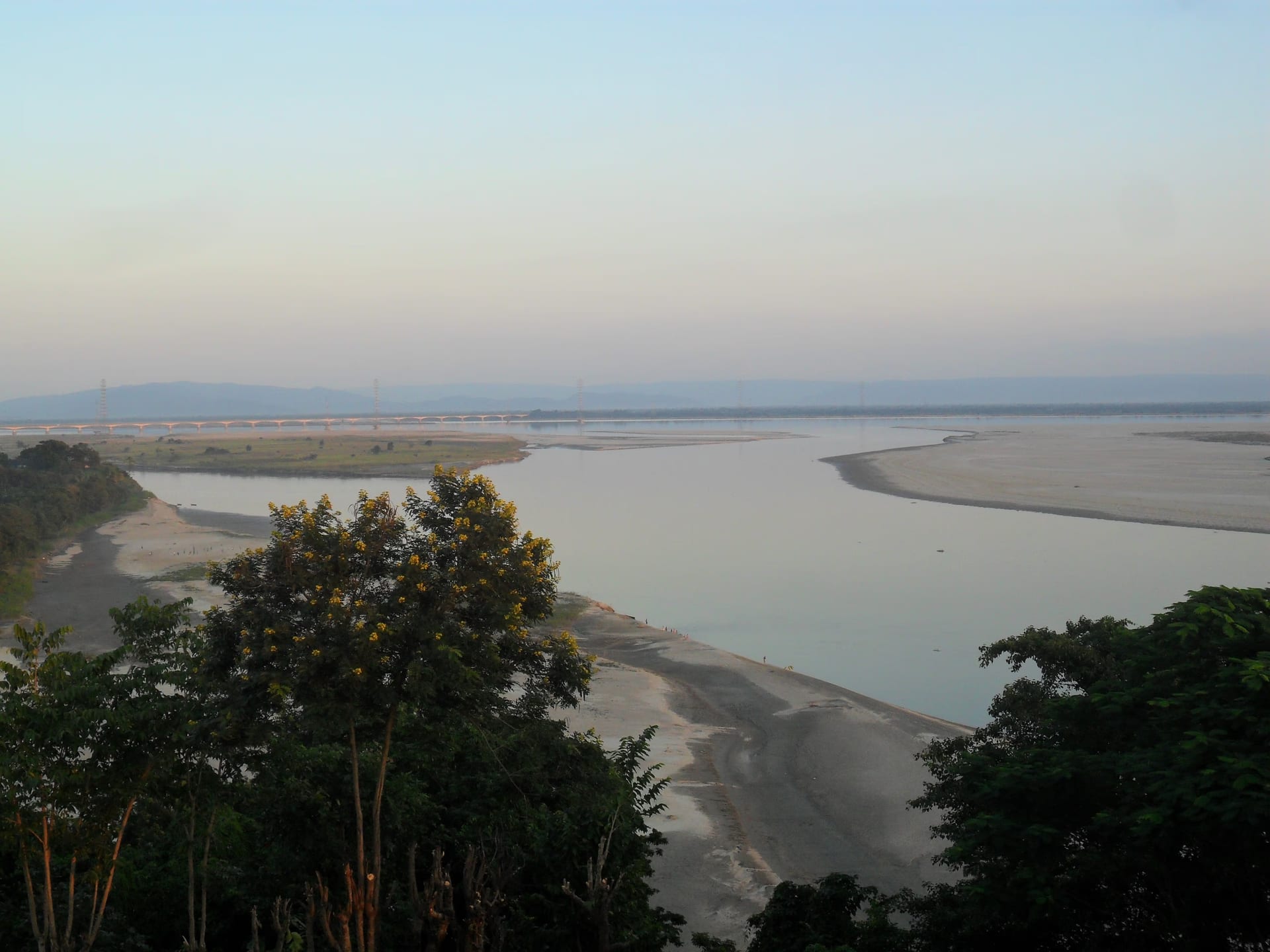 The Brahmaputra seen at dusk from the summit of Agnigarh hill at Tezpur, the river sweeping in a broad curve between pale sandbars with the long line of the Kolia Bhomora bridge crossing in the distance and low hills along the far bank