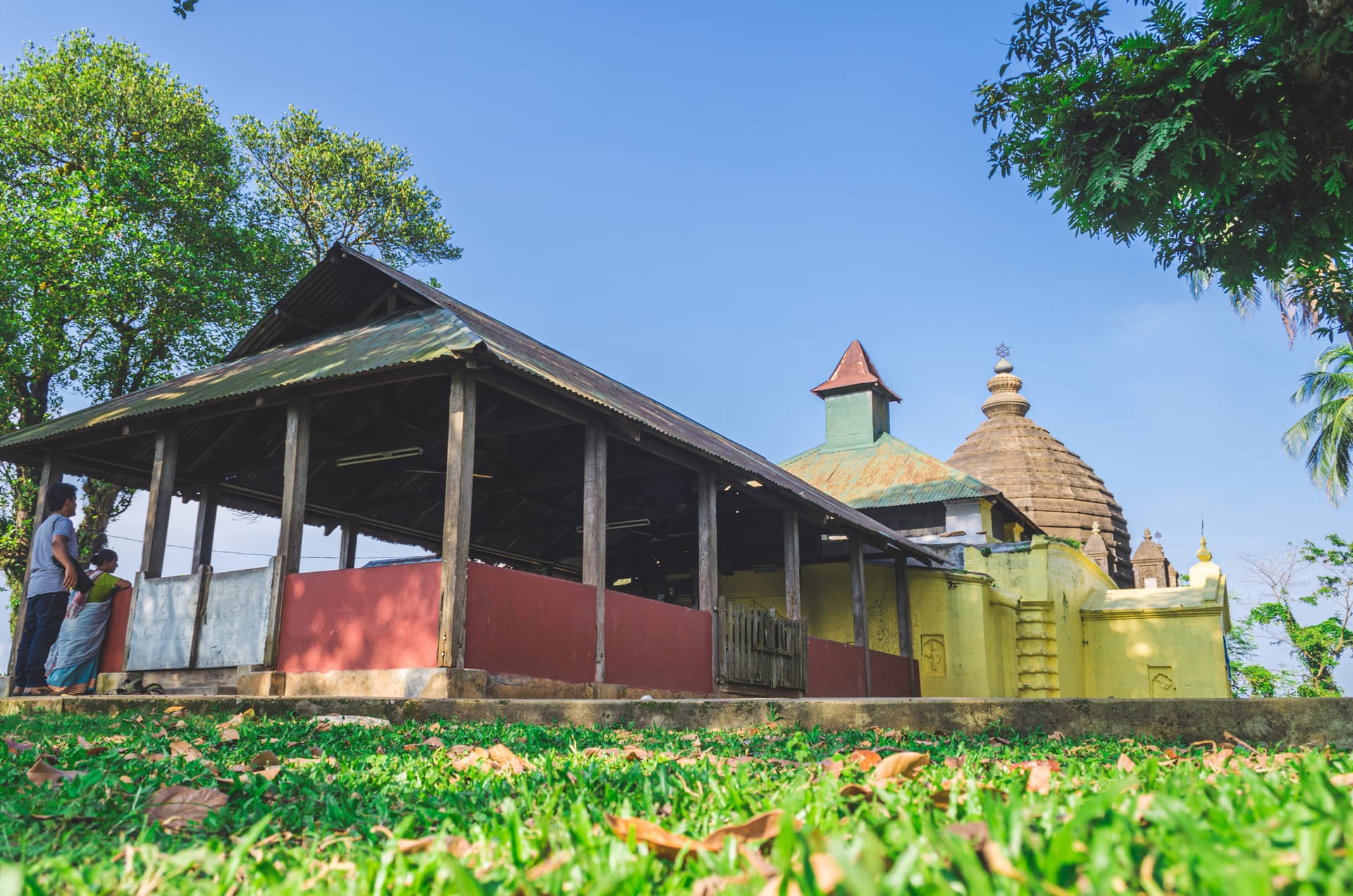 Side view of the Aswaklanta temple complex: a long tin-roofed open mandapa on stout wooden posts with a red-painted base, beside the yellow shrine with its conical metal tower and, behind it, the ribbed grey sikhara of the second shrine, under a bright blue sky framed by trees
