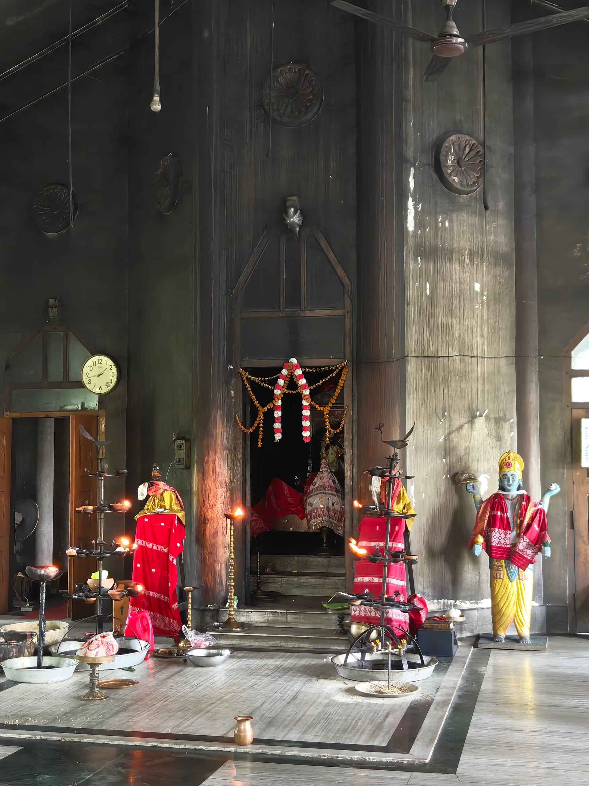 A high-ceilinged wooden shrine hall with a garlanded sanctum doorway, a painted deity figure, ceremonial standards and fly-whisks, and rows of brass offering lamps and vessels on the floor