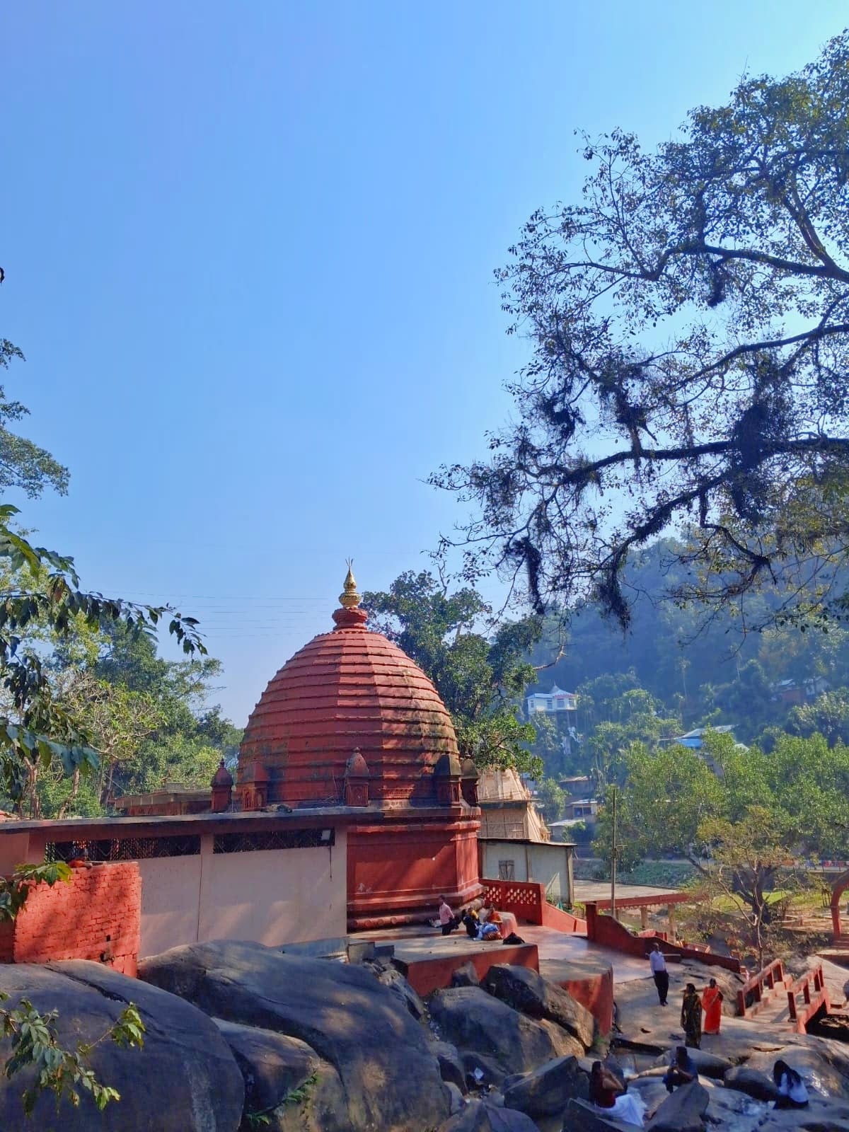 The Basistha temple complex with its ribbed orange-red sikhara set among large boulders, with forested hills rising behind under a clear blue sky
