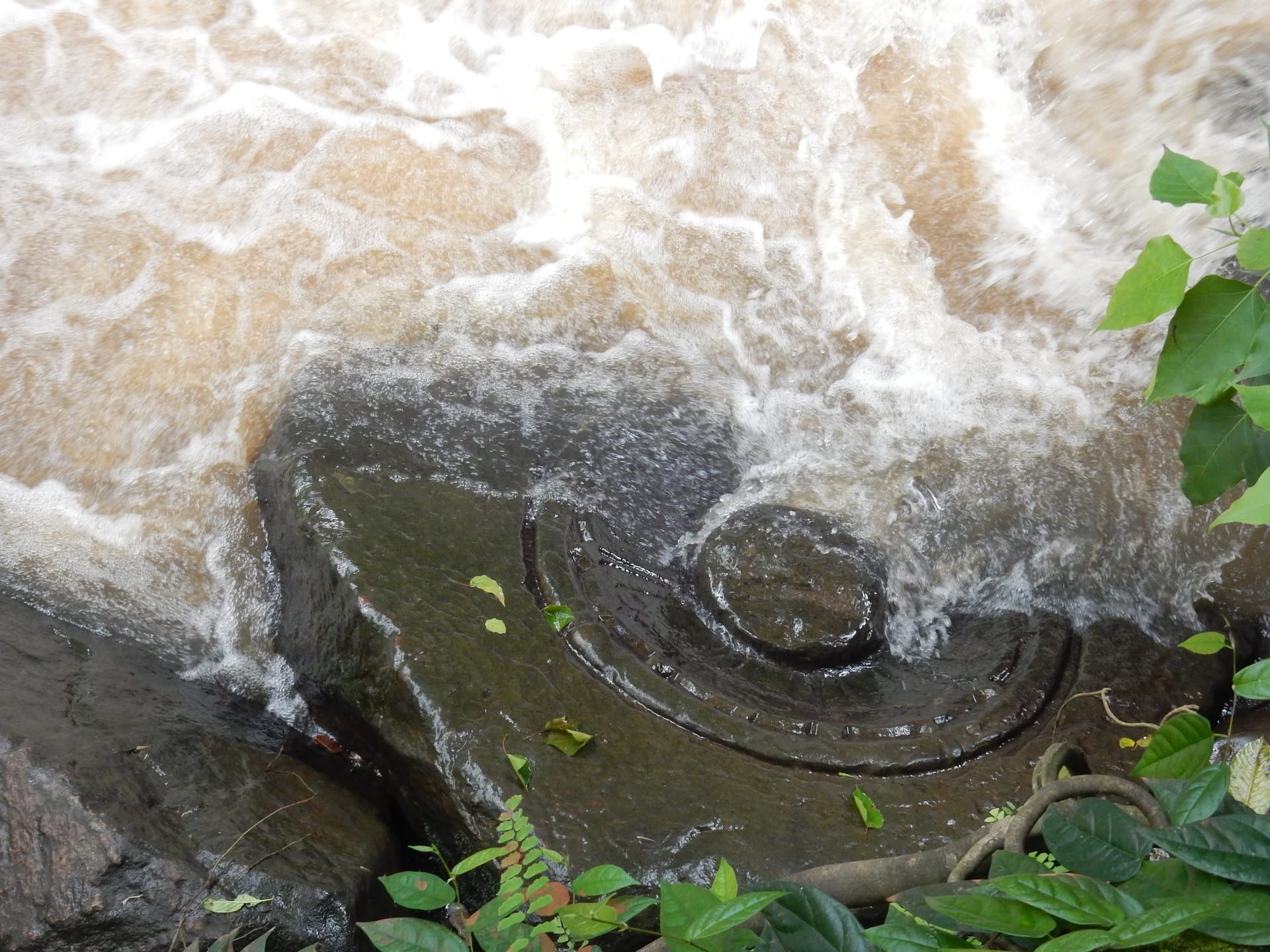 A Shiva lingam and its circular yoni carved into the dark bedrock of a stream, with white water rushing over it