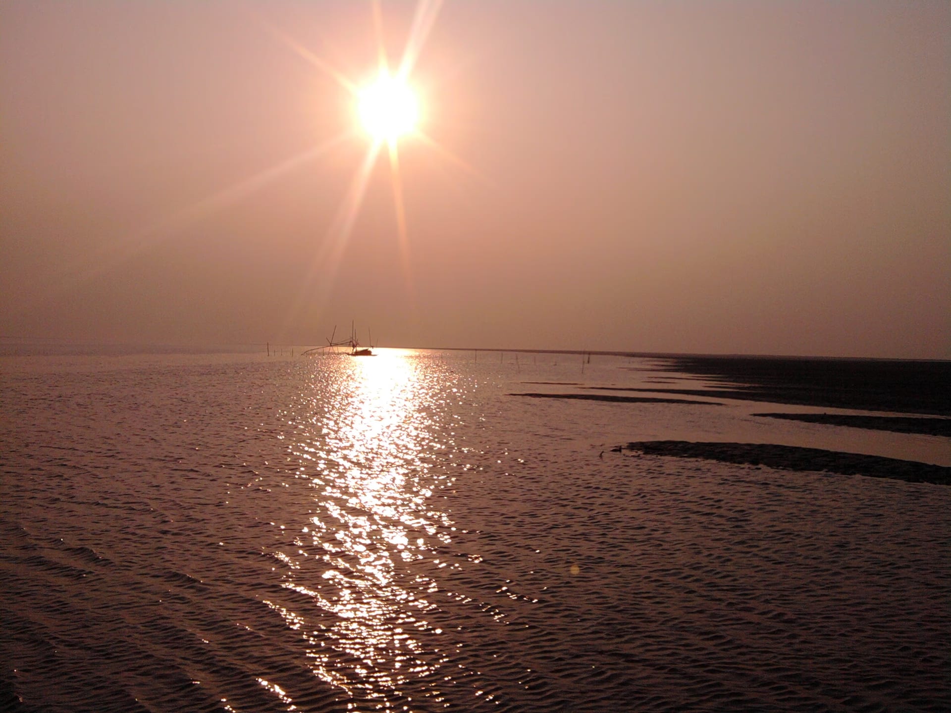 The sun low over the Brahmaputra at Biswanath Ghat, light tracking across the water past a small fishing boat to exposed sandbanks on the right